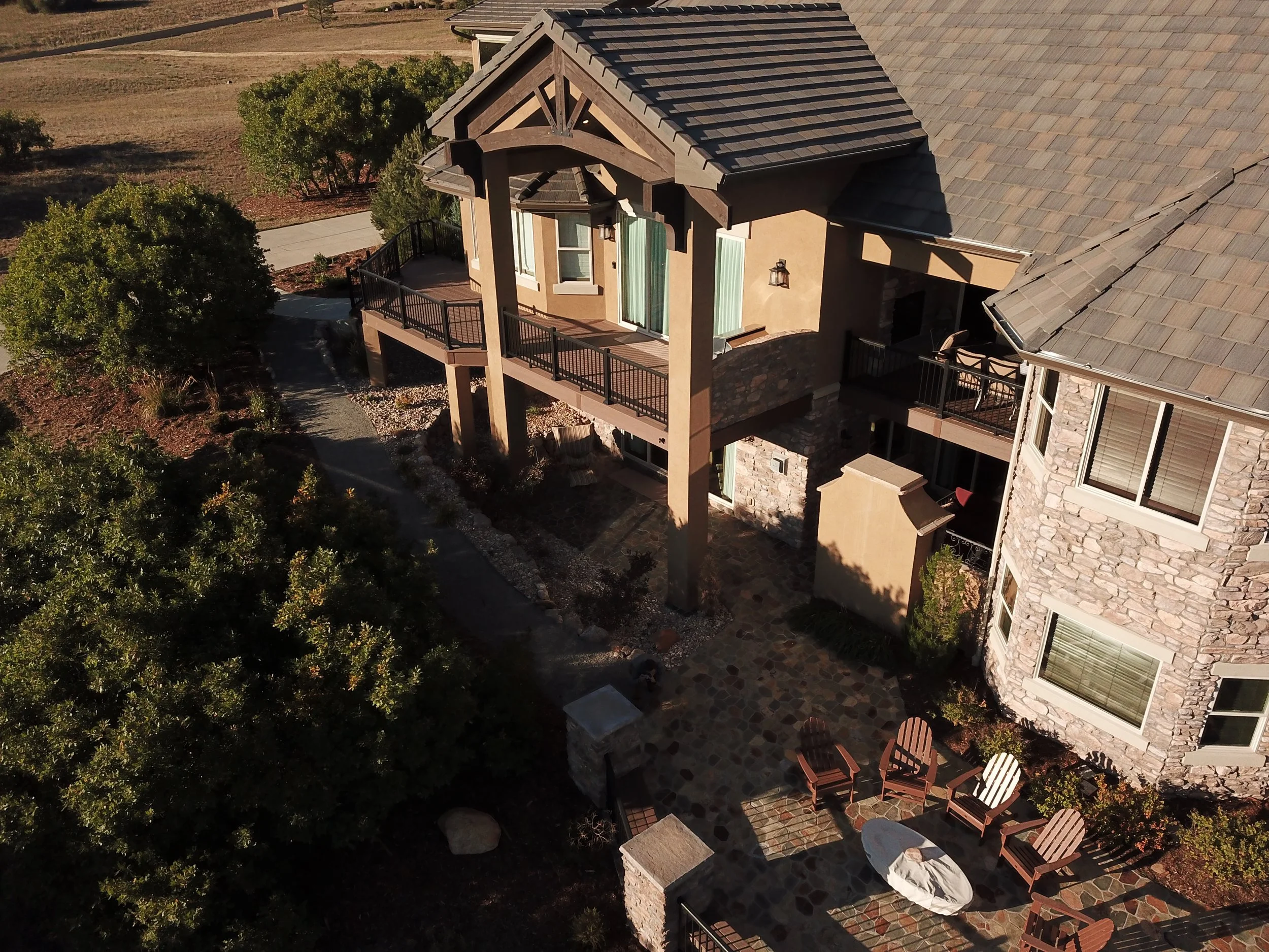 Aerial view of a two-story house with a patio area featuring outdoor chairs and a fire pit, surrounded by trees and near an open field.