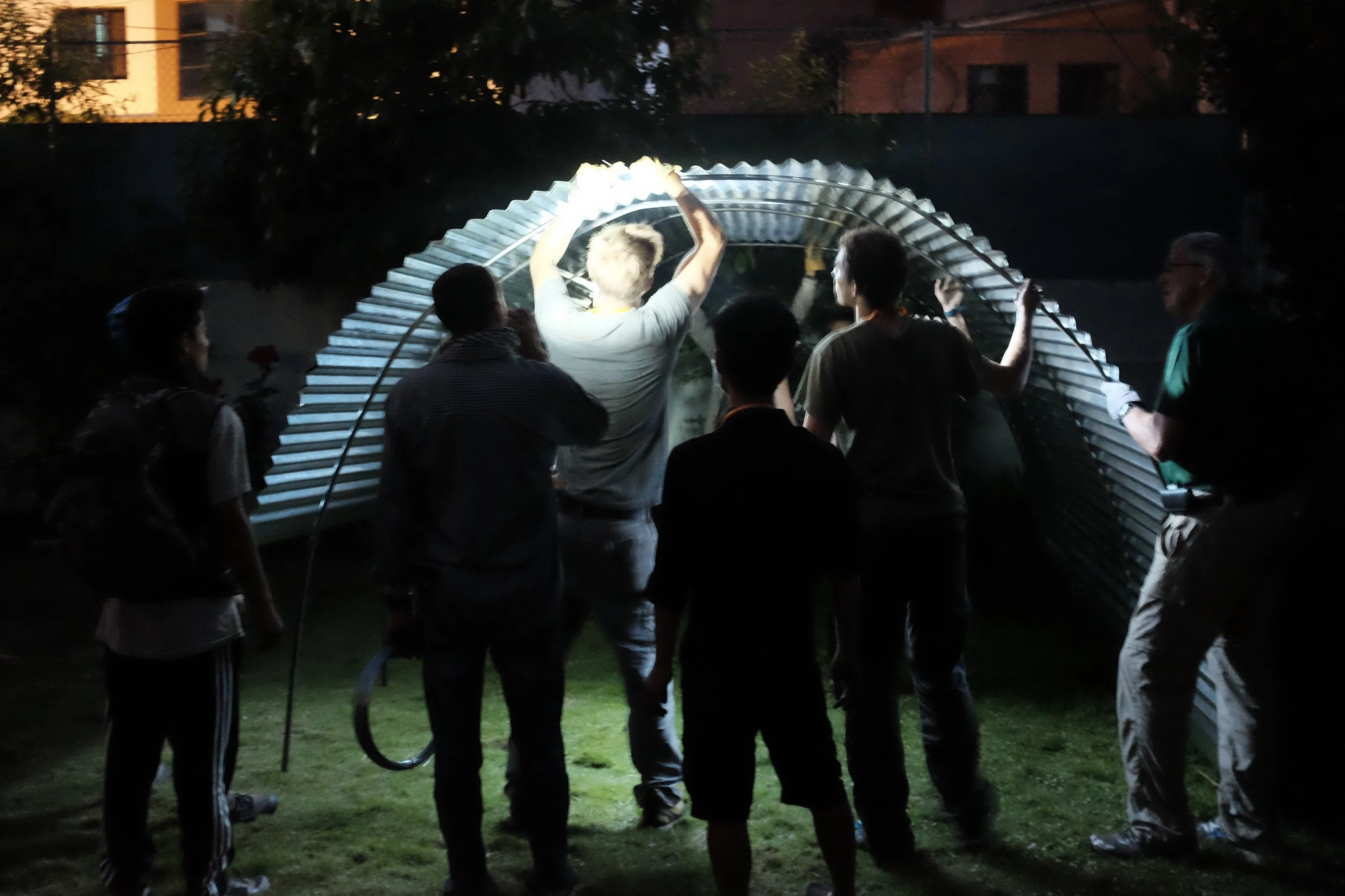 Group of people working together to assemble a large arch-shaped structure outdoors at night, illuminated by a nearby light.