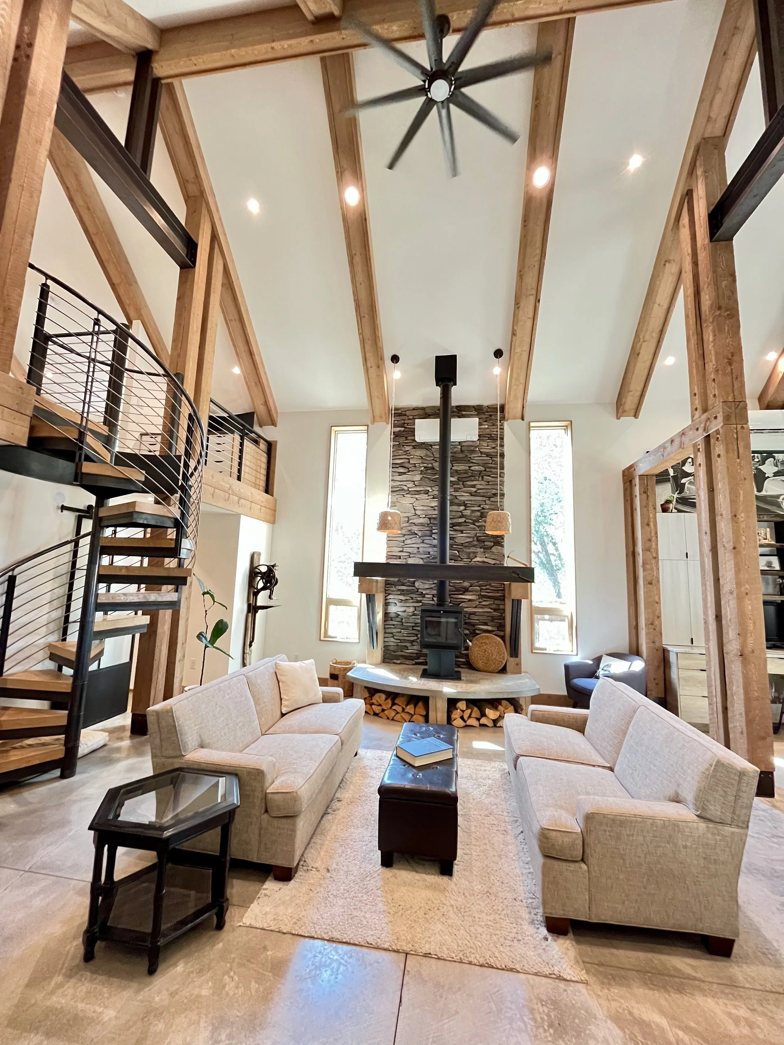 Living room with high vaulted ceiling and exposed wooden beams, stone fireplace with wood storage underneath, beige sofas, a small black side table, a leather ottoman, and large windows allowing natural light.