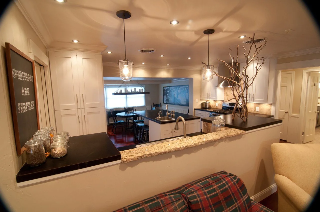 View of a kitchen with white cabinets, black countertops, and modern pendant lights hanging from the ceiling. There is a central island with a sink, a large branch centerpiece, and jars on the counter. In the background, there is a dining area with a
