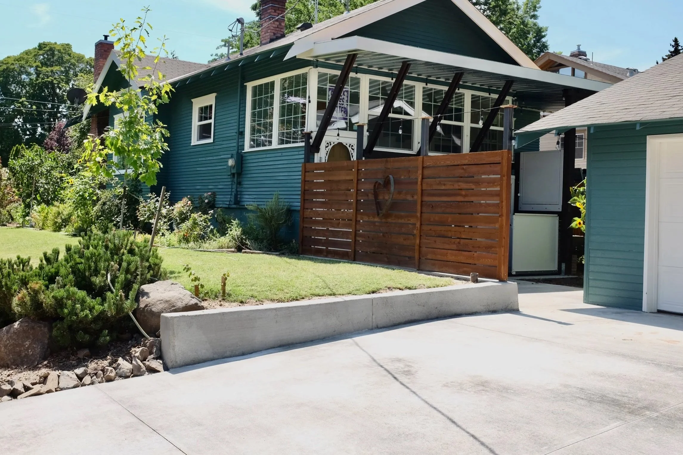 Backyard of a blue house with a small garden, a concrete driveway, a wooden privacy fence with a heart-shaped decoration, and a covered porch area.
