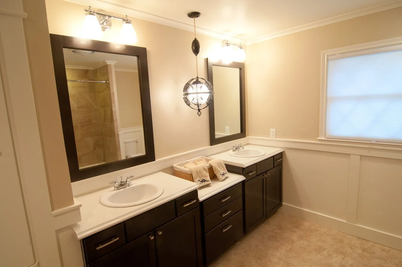 Bathroom with two dark wooden vanities, each with a white countertop and oval sinks. Two large rectangular mirrors are above the vanities. There are three light fixtures above each mirror, and a decorative hanging light in the center. The wall is pai