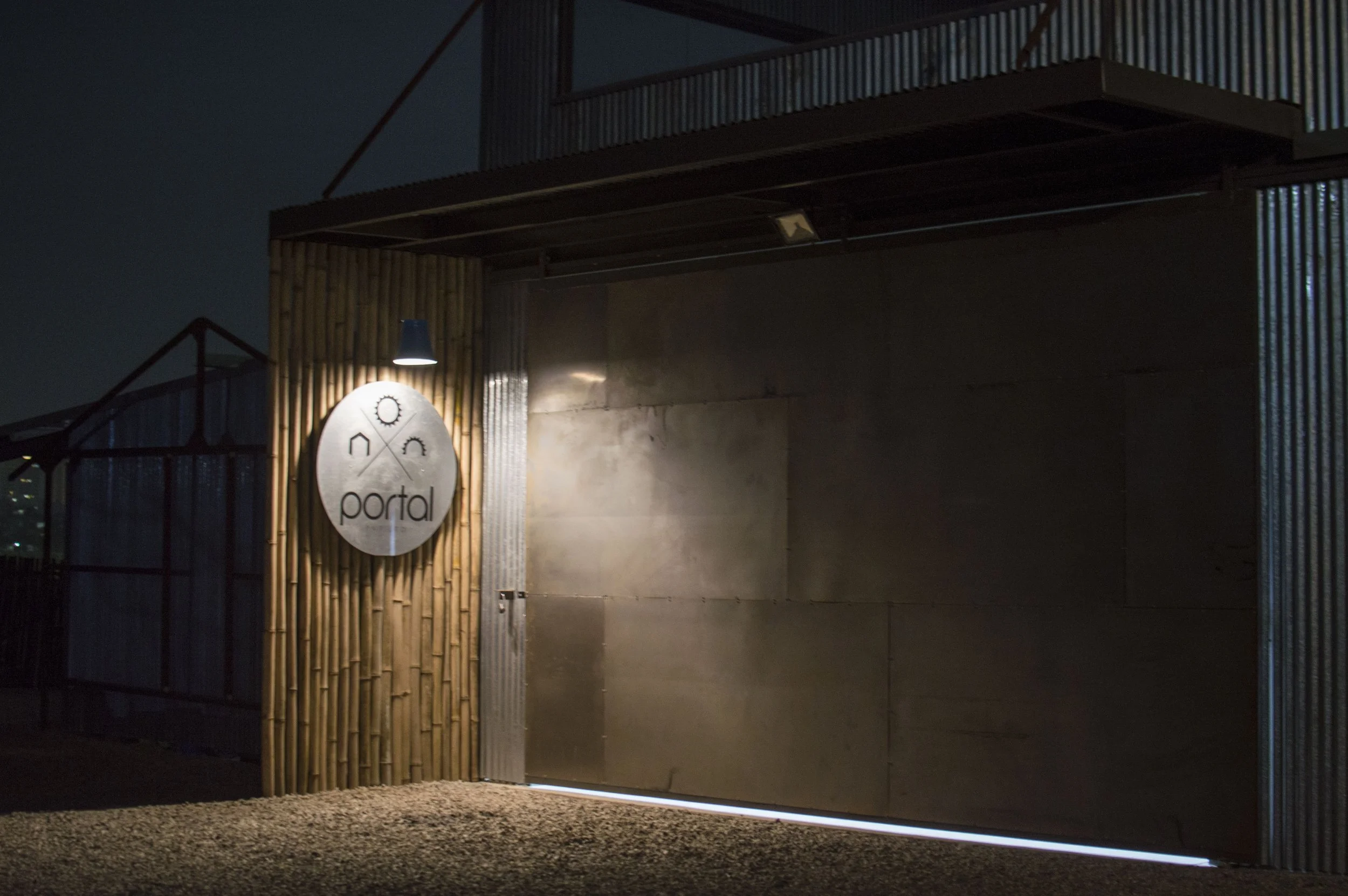 Nighttime photo of a modern building entrance with a round sign that reads 'portal' and has icons of a house, a gear, and a sun. The entrance features corrugated metal and wooden cladding, illuminated by a small overhead light.