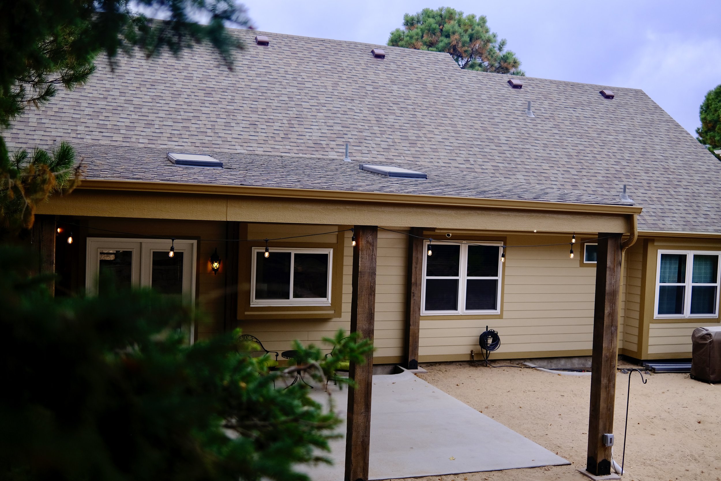 Back of a house with a beige exterior, brown roof with skylights, string lights hanging, and a concrete patio with sand and utility hookup.
