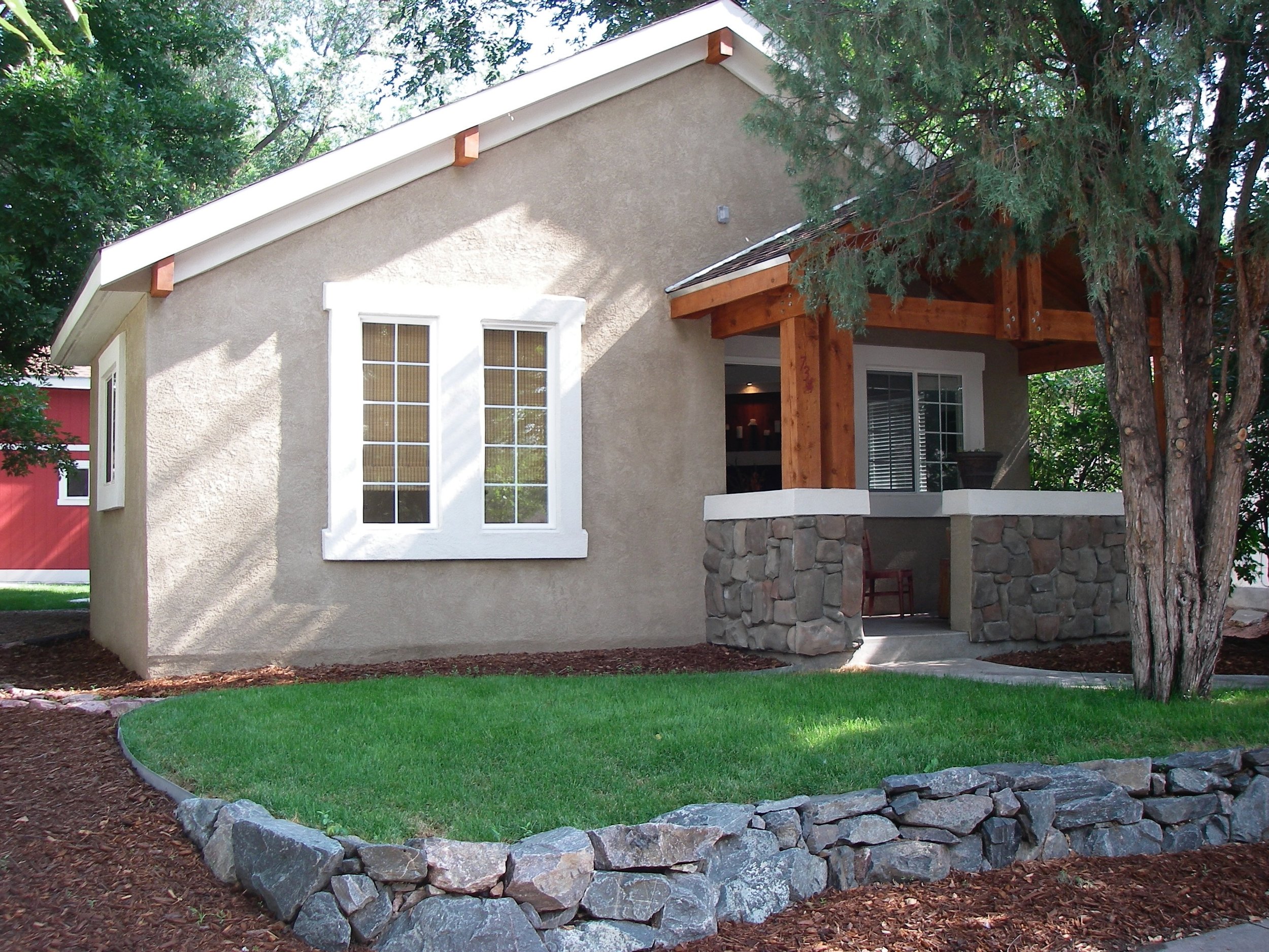 A small house with beige stucco walls and a gabled roof. The house has white trim around the windows and a porch with stone columns and wooden support beams. There is a well-maintained lawn and a stone pathway leading to the porch, with trees and lan