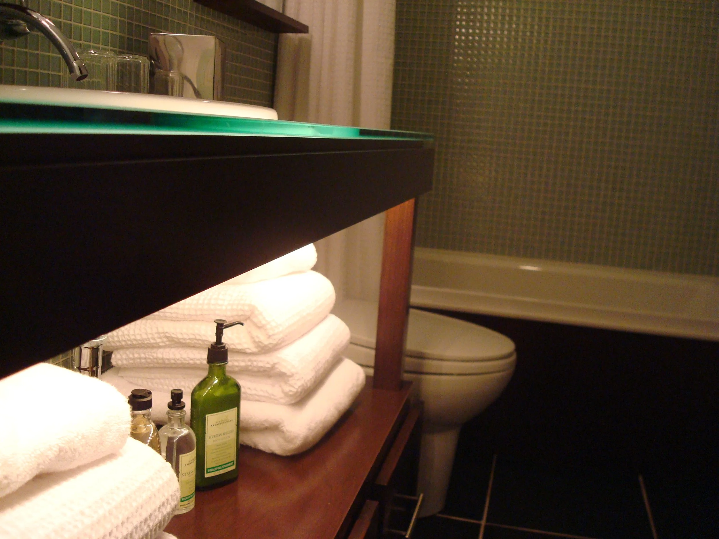 A bathroom vanity with stacked white towels, small bottles of toiletries, a sink, a faucet, and a mirror. In the background, a toilet is visible next to the shower with a tiled wall.