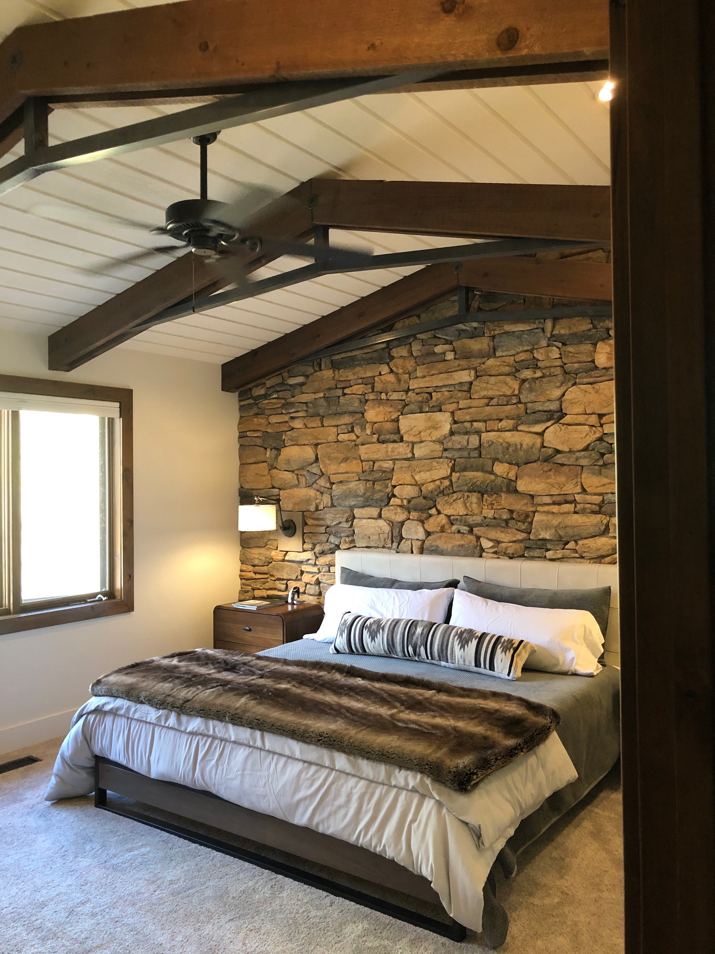 A cozy bedroom featuring a stone accent wall behind a large bed with white and gray bedding, a faux fur throw blanket, and striped decorative pillow. There's a wooden nightstand and wall-mounted lamp, a window with wooden trim, and exposed wooden cei