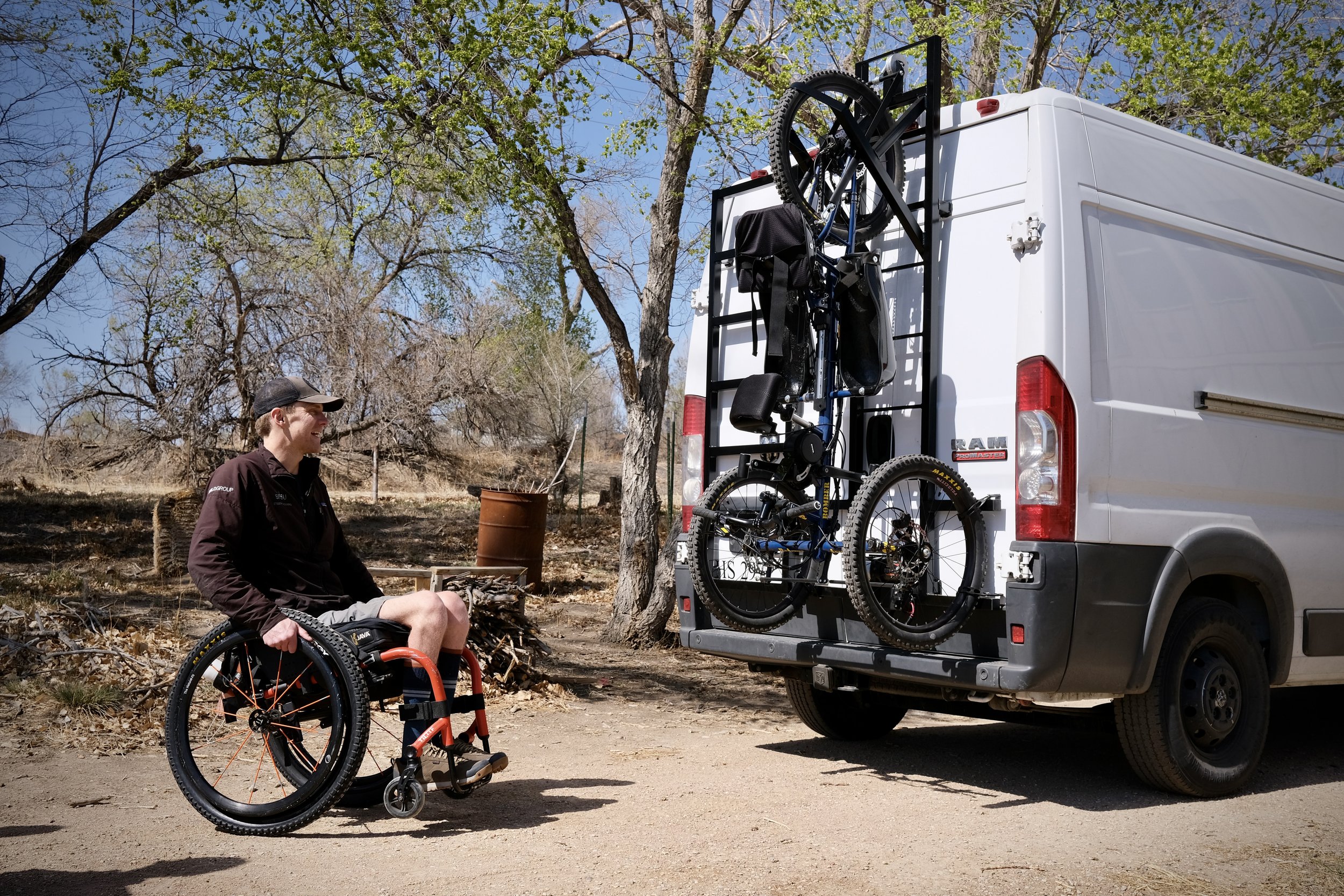 A person in a wheelchair and a van with a mounted bicycle rack holding two bicycles outdoors near trees and dry grass.
