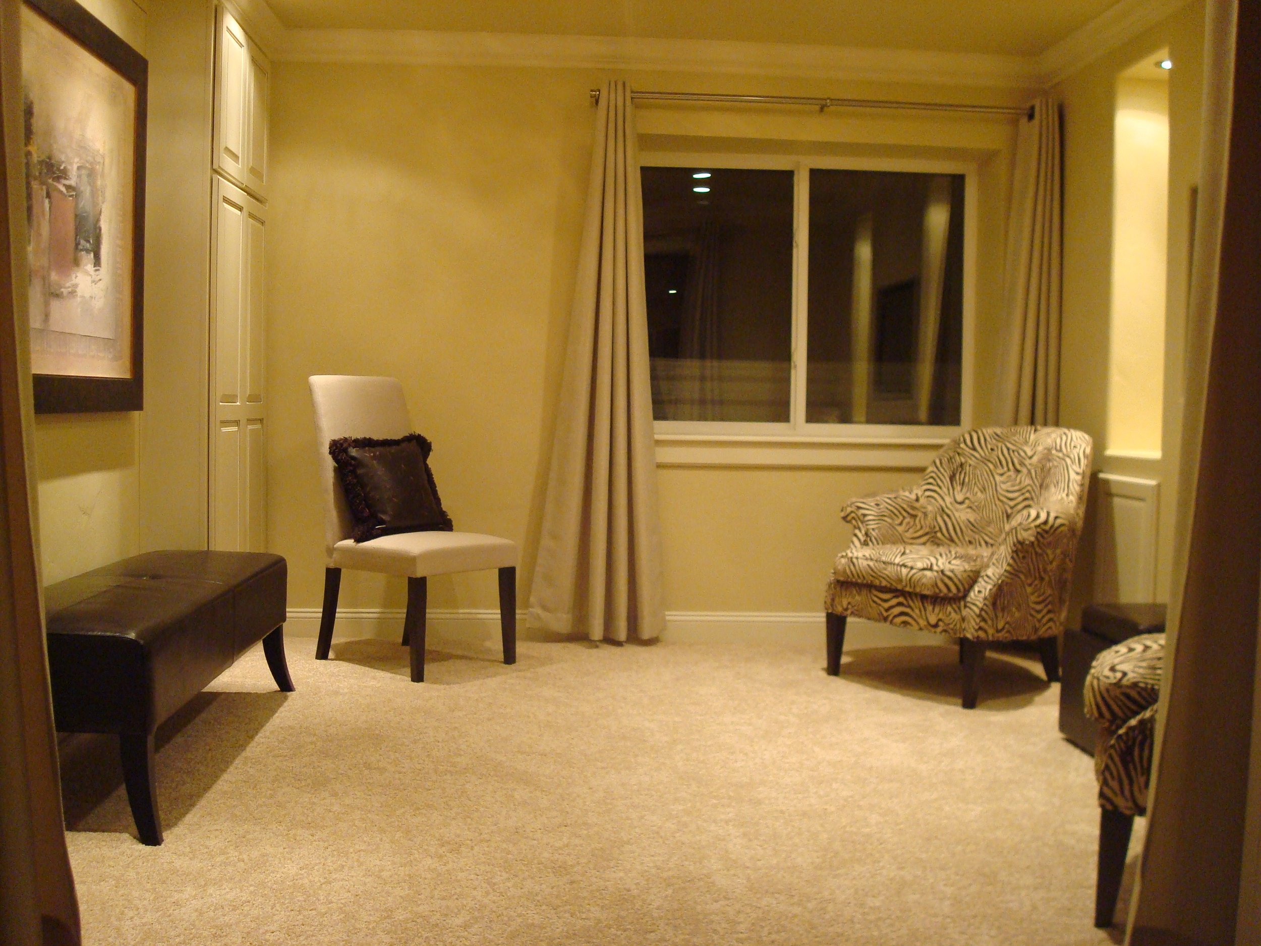 A cozy living room with beige walls and carpet, featuring a zebra-print armchair, a white upholstered chair with a dark cushion, a dark cushioned bench, and a window with cream curtains at night.