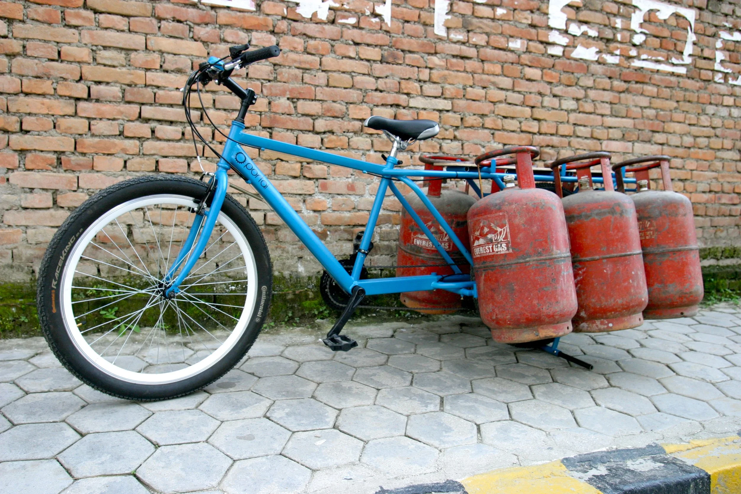 A blue bicycle converted to transport four propane gas tanks, parked on a sidewalk against a brick wall.