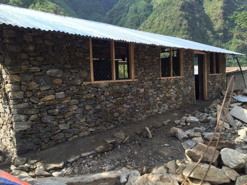 Stone house under construction with a metal roof, surrounded by construction debris and mountains in the background.
