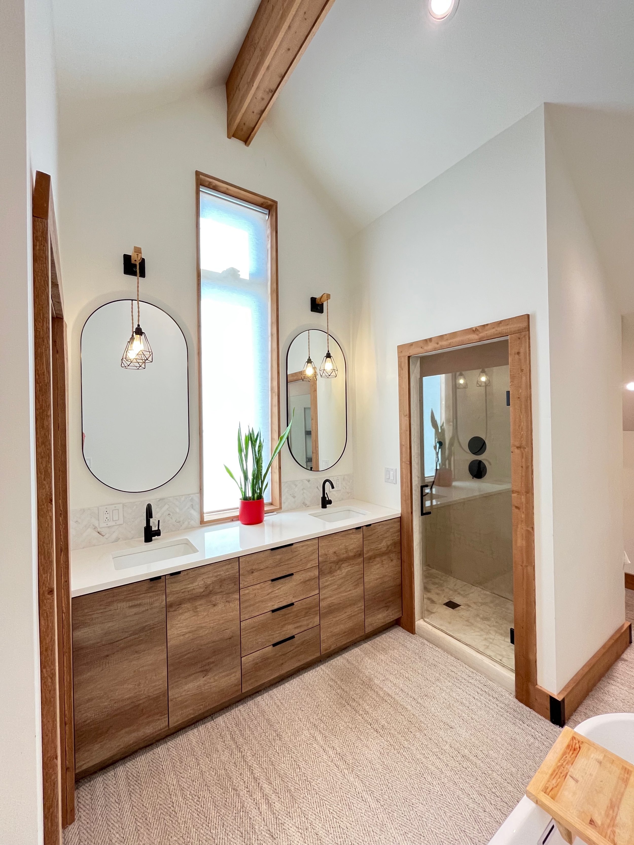 A modern bathroom with a double vanity, two oval mirrors, two pendant lights, a plant in a red pot, and a glass-enclosed shower with wood trim.