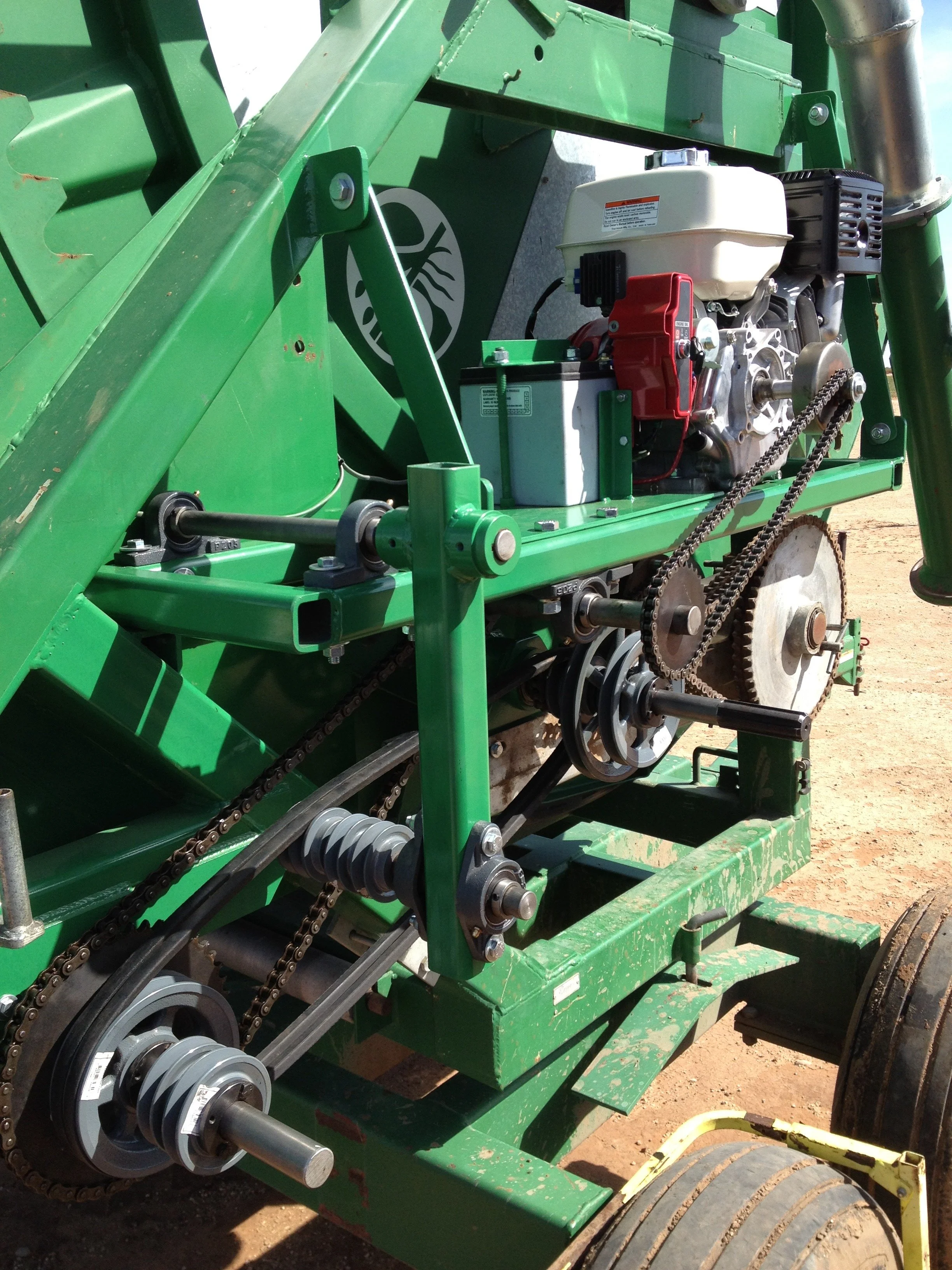 Close-up of green agricultural machinery with chains, gears, and engine components, on a dirt surface.