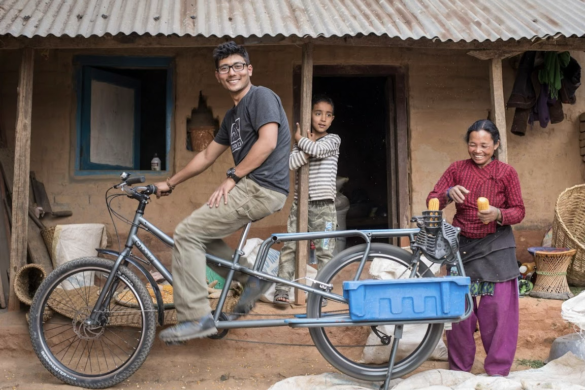 A young man rides a bicycle in front of a rural house, with a woman holding some snacks and two children standing in the doorway, all smiling.