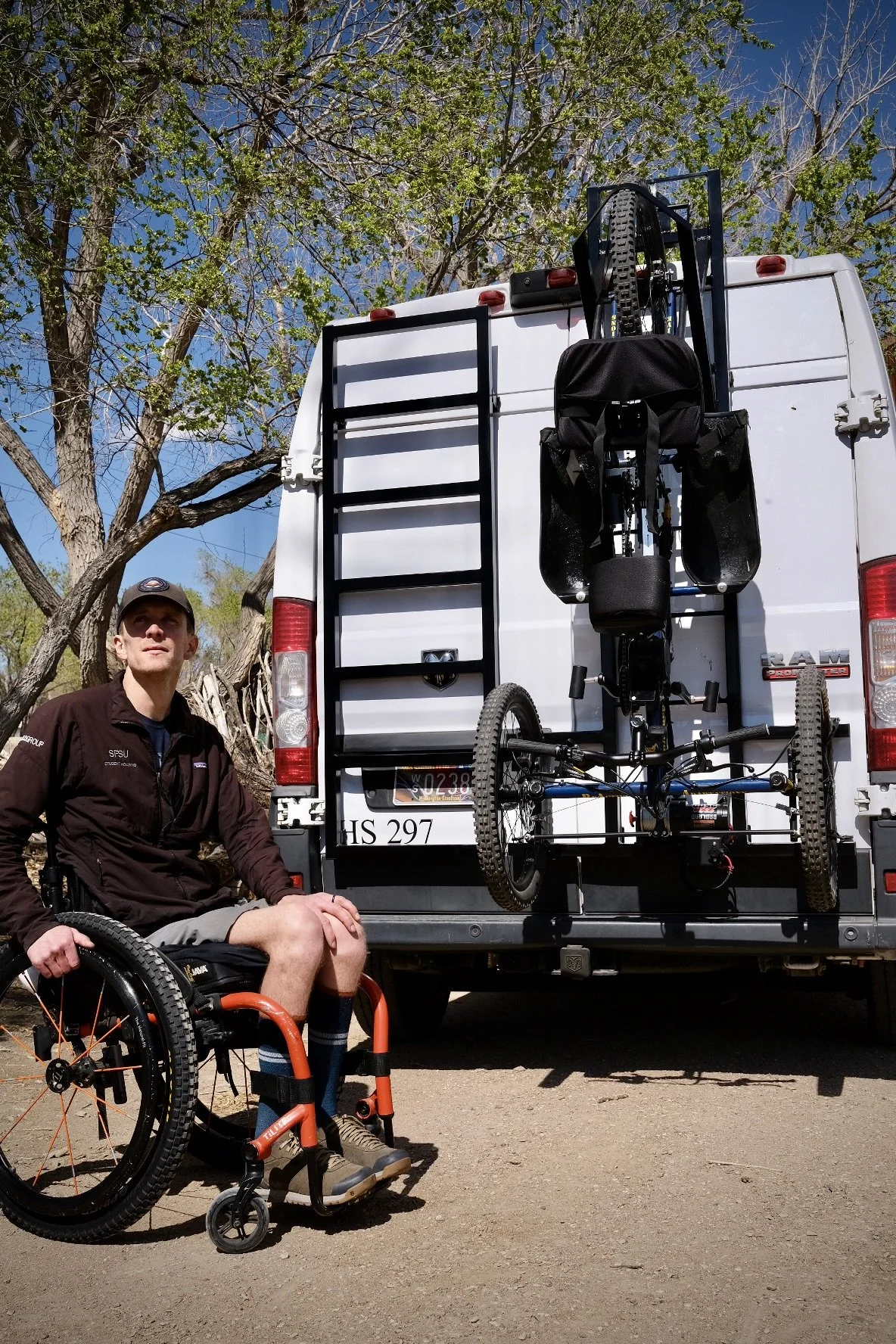 A man in a wheelchair sits next to a van equipped with a bike rack carrying two bikes. The man is wearing a black jacket and a cap, and the background shows trees and a clear blue sky.