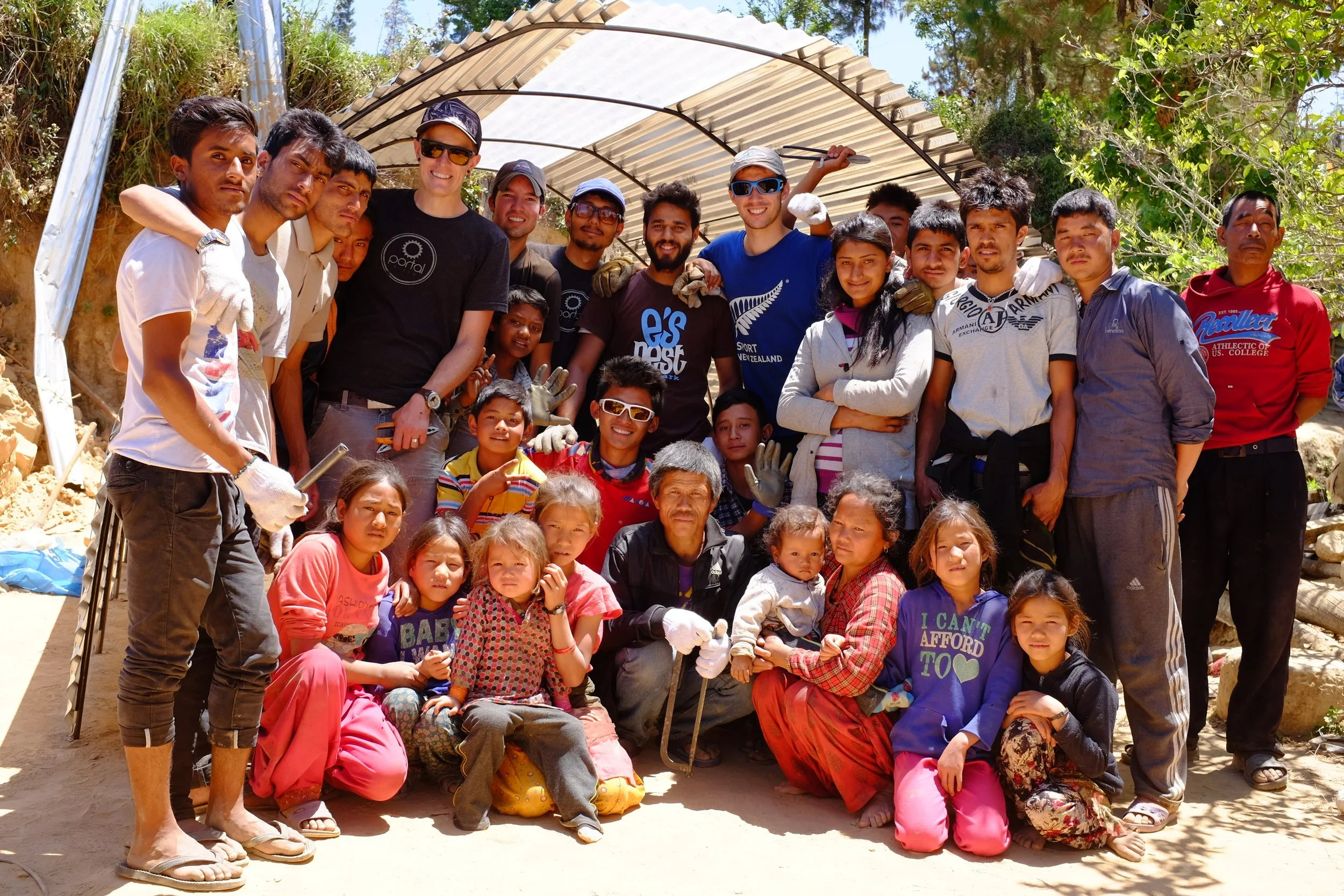 A large group of people, including children and adults, posing together outdoors under a partial shelter with trees and rocks in the background. The group appears happy, and some are wearing gloves.