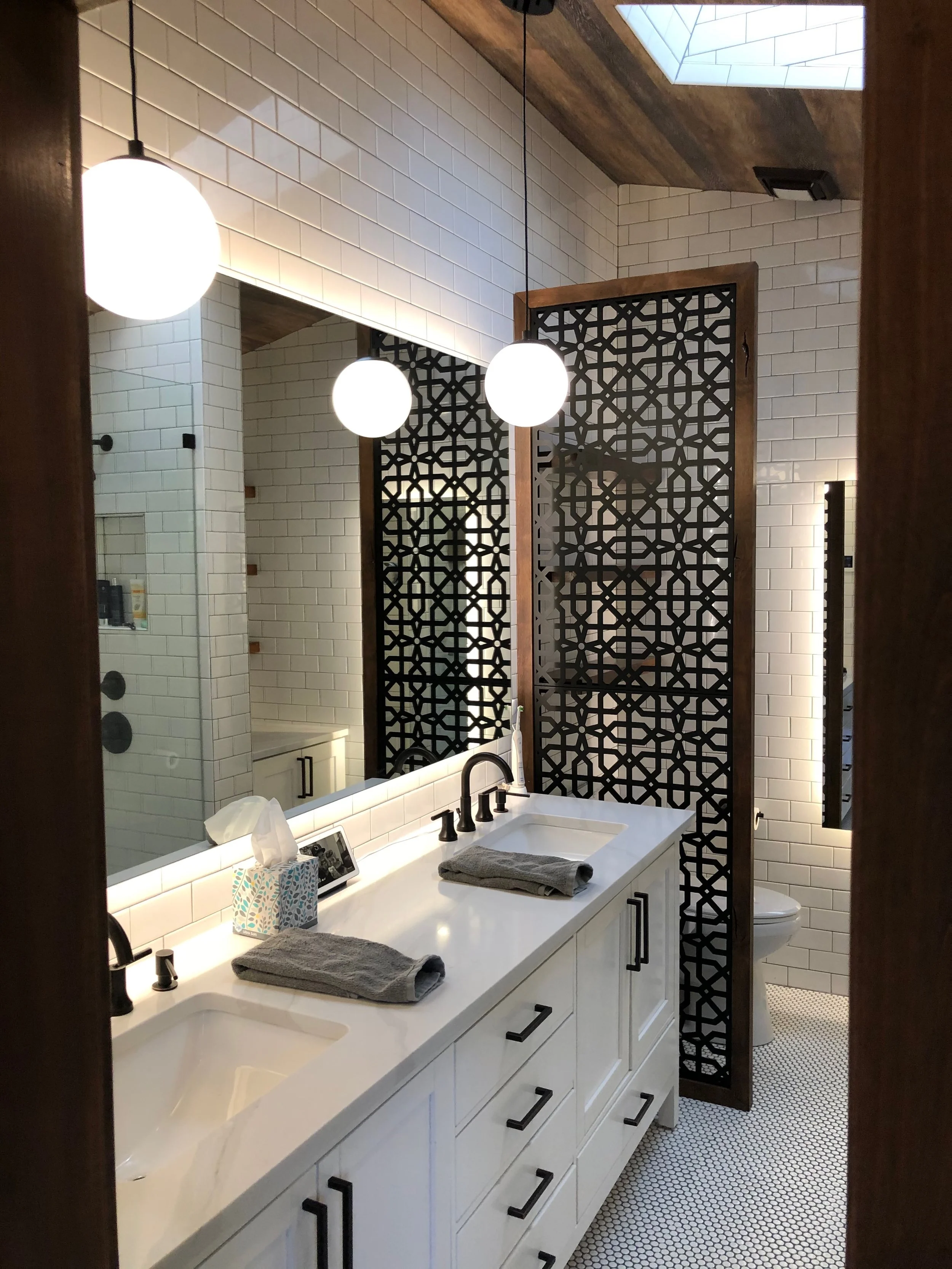 Modern bathroom with white tiled walls, black fixtures, double vanity with a white countertop, two sinks with towels, large mirror, geometric wooden privacy screen, black pendant lights, and a skylight.