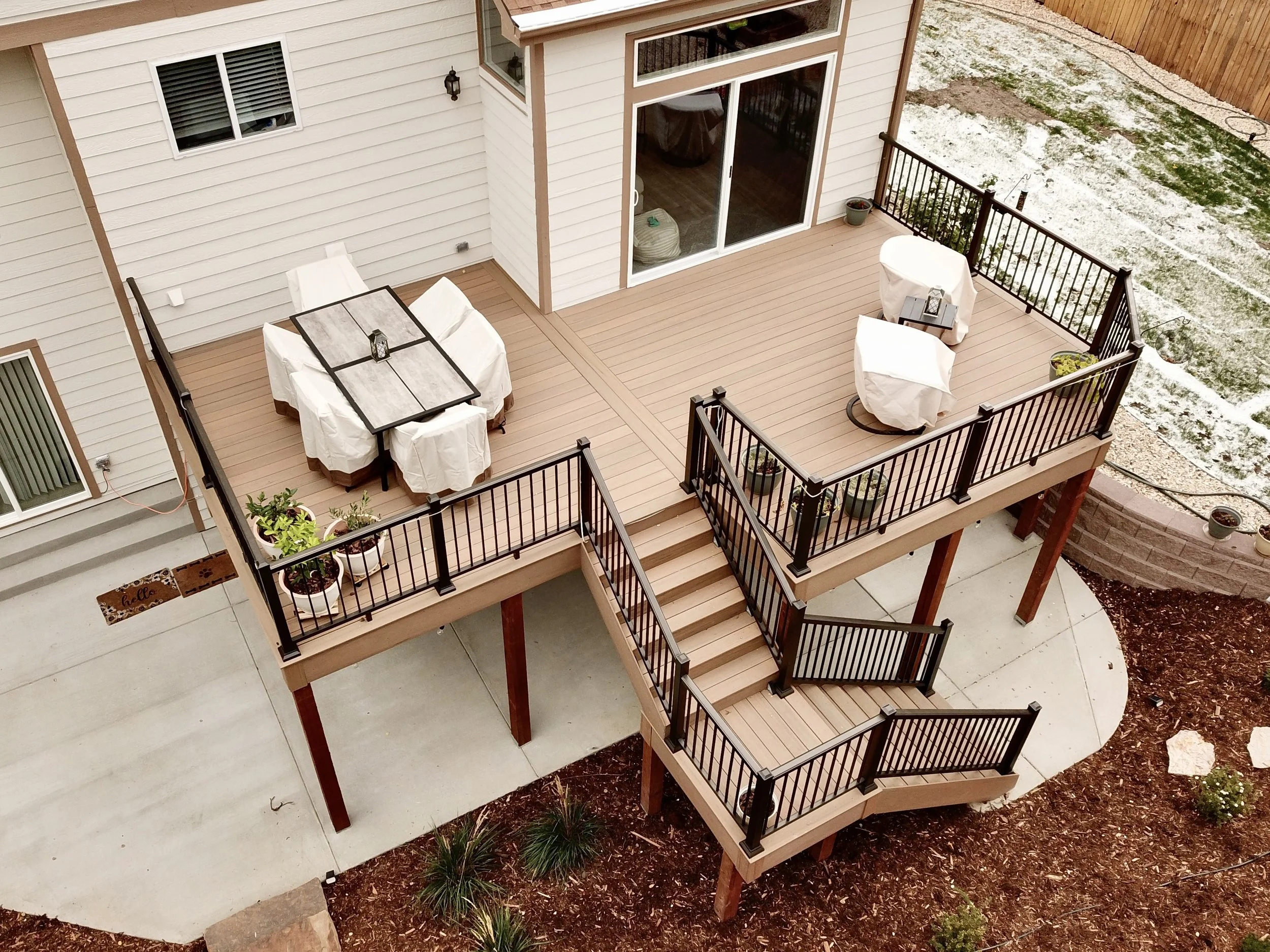 A backyard deck with a dining table covered in a white cloth, six chairs also covered in white cloth, and two round chairs with white covers. There are potted plants on the deck and a small table with a lantern and a book. The deck has stairs leading