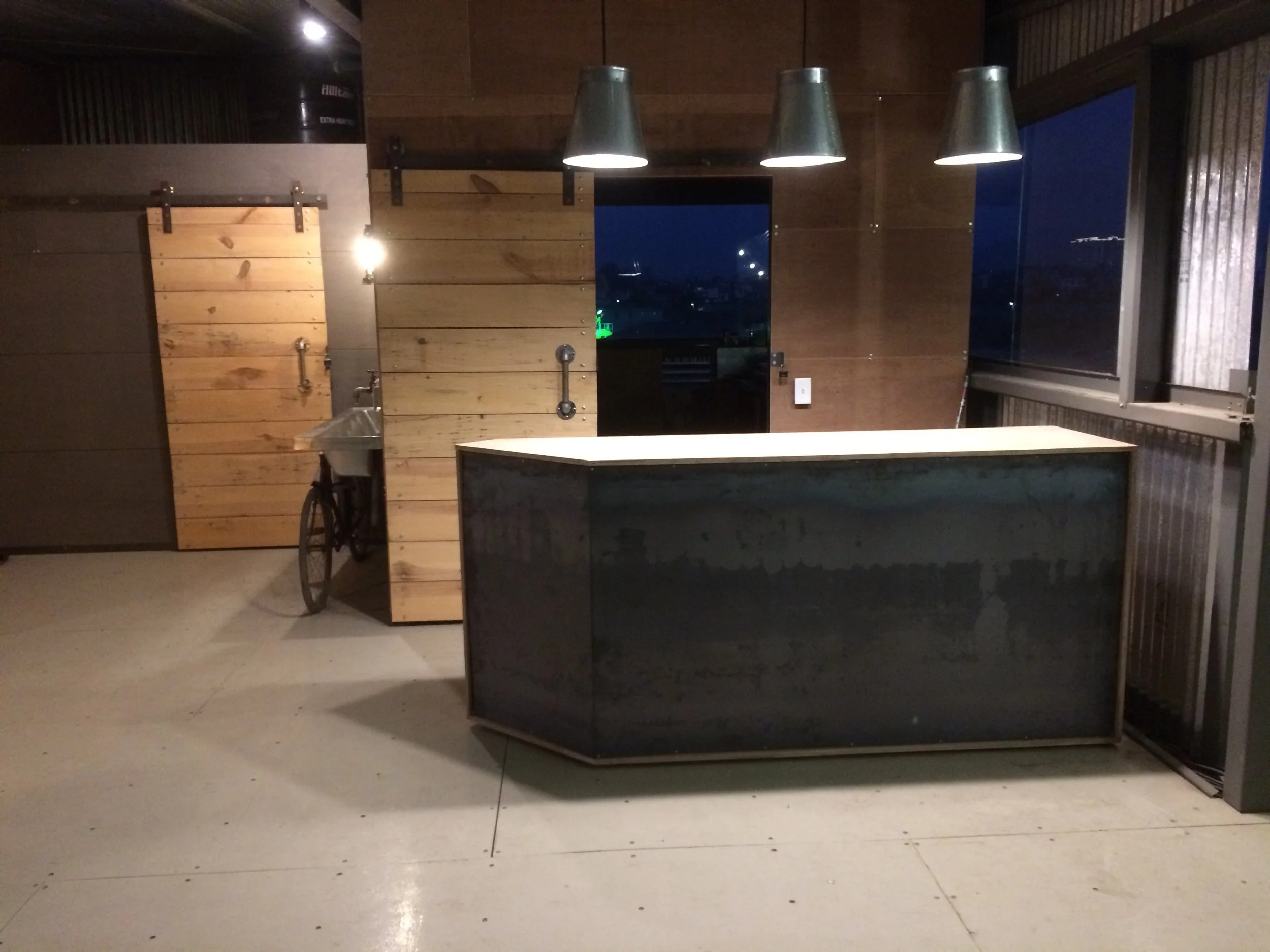 Minimalist kitchen with a black marble island, three metallic pendant lights, wood panels on the wall, a small rustic sink with a wheel, and a window showing a night city view.