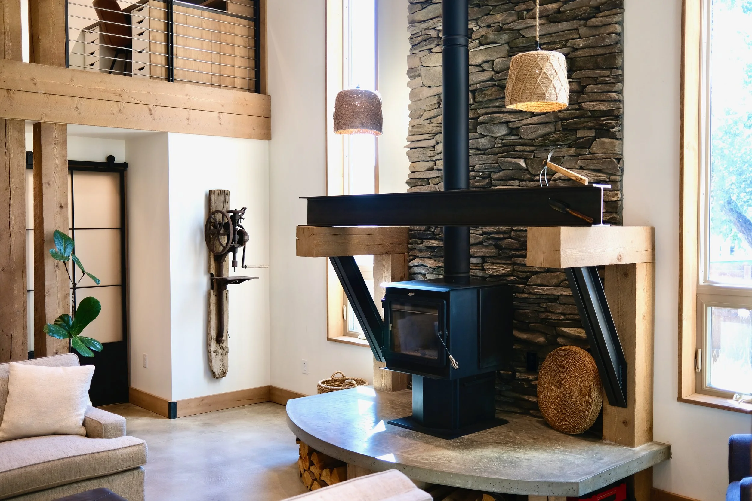 Living room with wood and stone accents, a black wood stove, wrapped in wood panels, with hanging woven light fixtures.