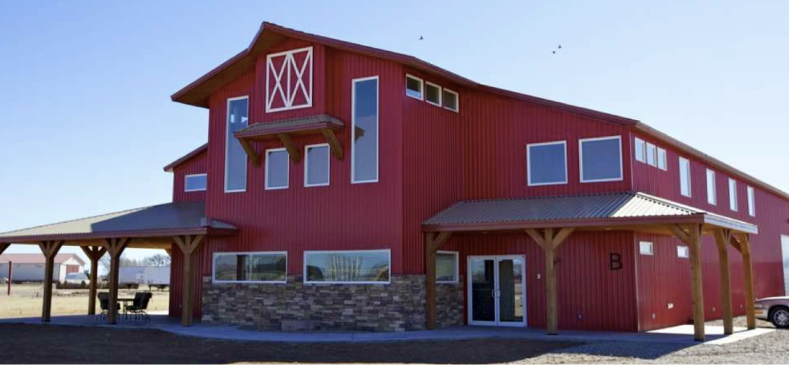 Large red barn-style building with multiple windows, stone accents on the lower part, and a covered porch with wooden supports, set against a clear blue sky.