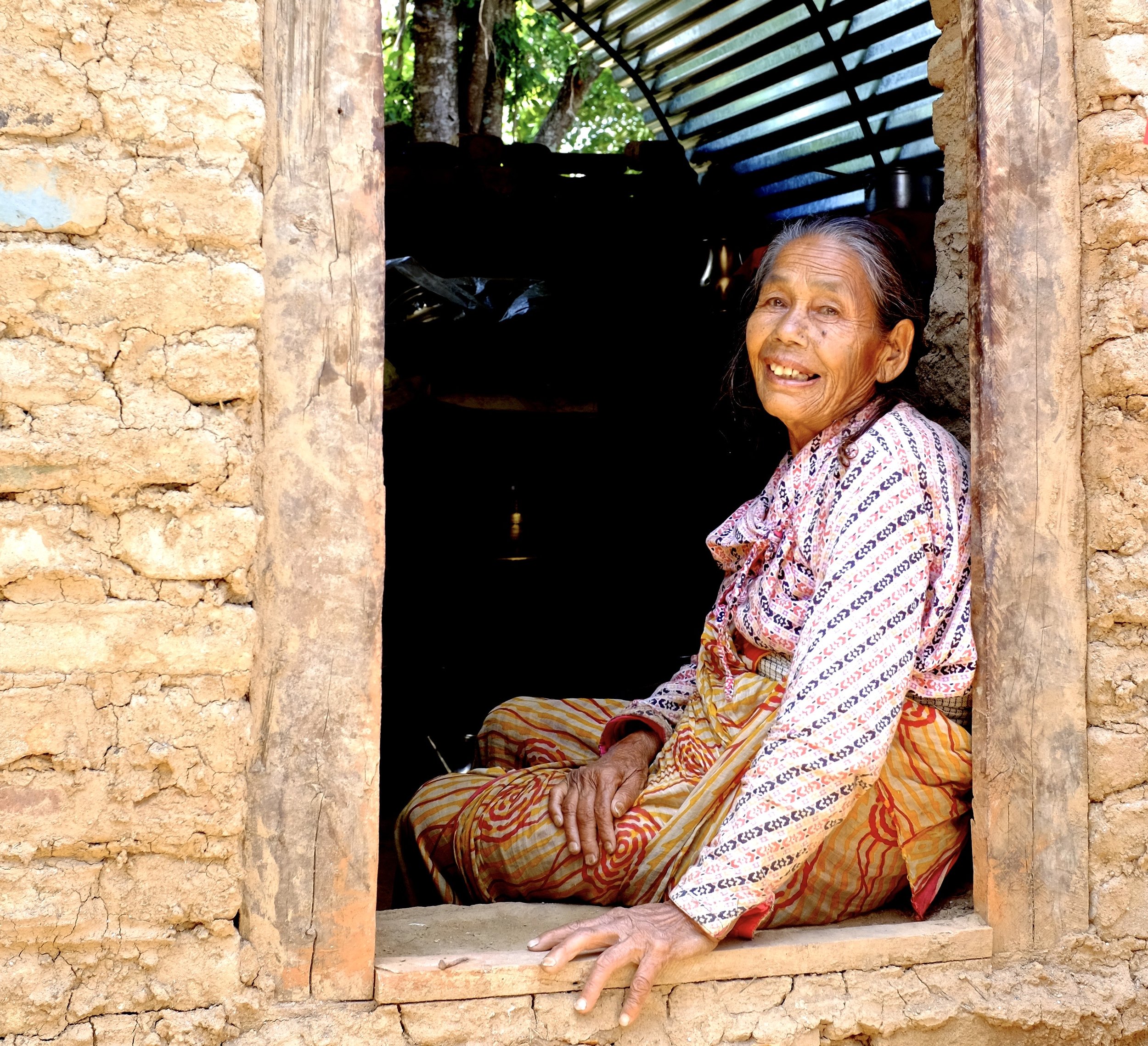 An elderly woman sits at a window in a rustic, mud-brick house, smiling warmly.