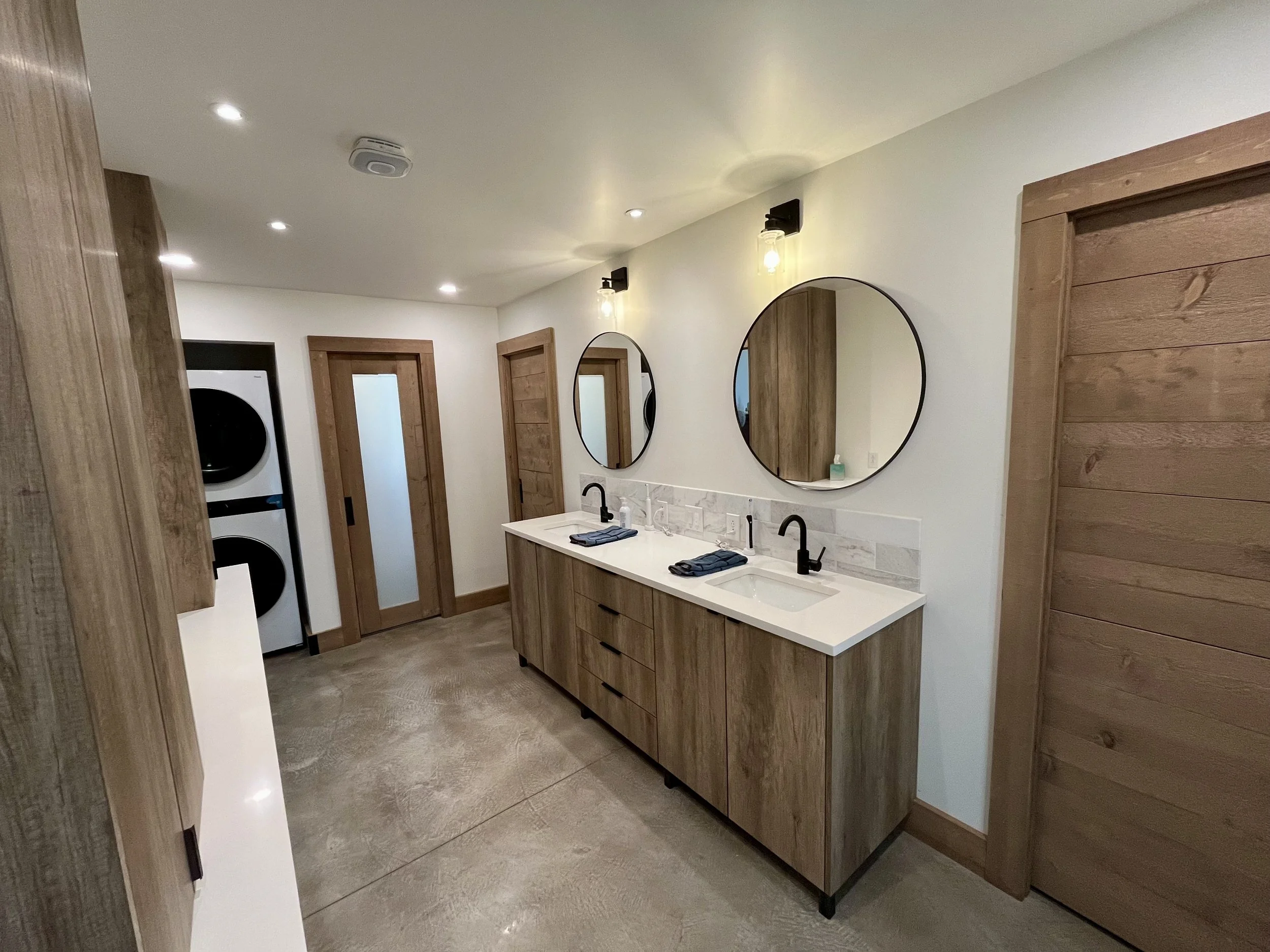 Modern bathroom with double vanity, two round mirrors, black fixtures, wood accents, and a laundry pair in the corner.
