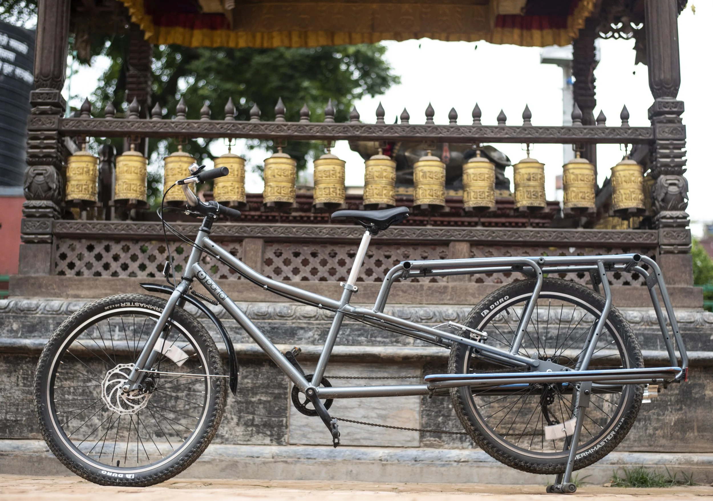 A silver cargo bicycle with a rear rack parked in front of a traditional Asian temple with ornate wooden carvings and gold prayer wheels.