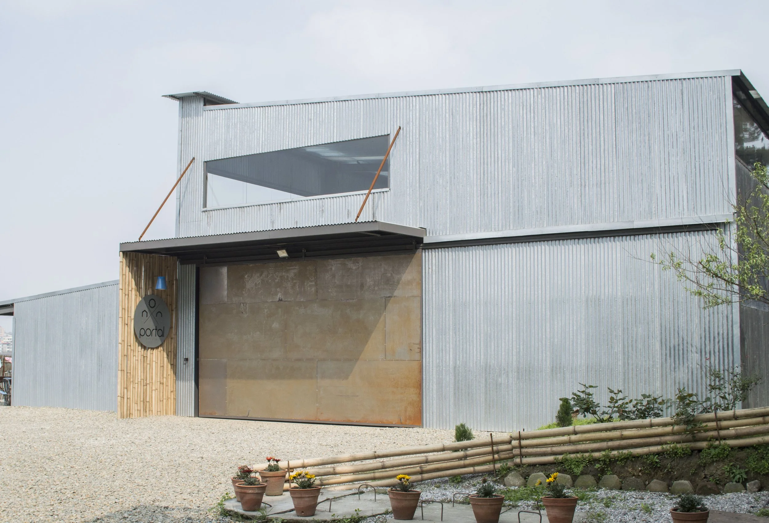 Modern building with corrugated metal walls, a large garage door with rusted metal panels, wooden accents, and a sign that reads 'portal' on a round black background. Potted plants and a gravel ground in front.