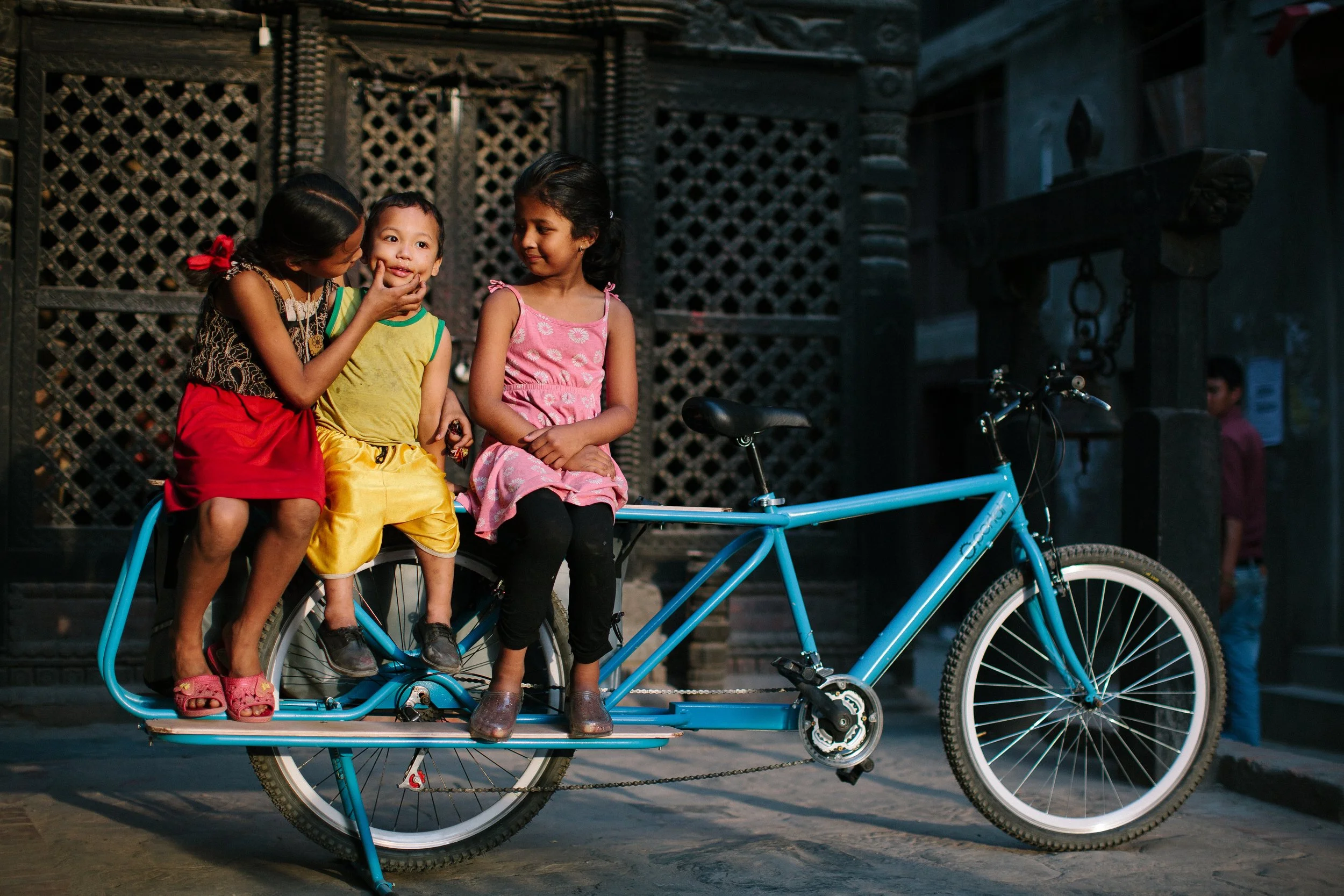 Three children sitting on a blue bicycle in front of an ornate, dark building. Two girls are wearing colorful dresses and a boy in yellow shorts. One girl is whispering into the boy's ear, with the other girl watching them.