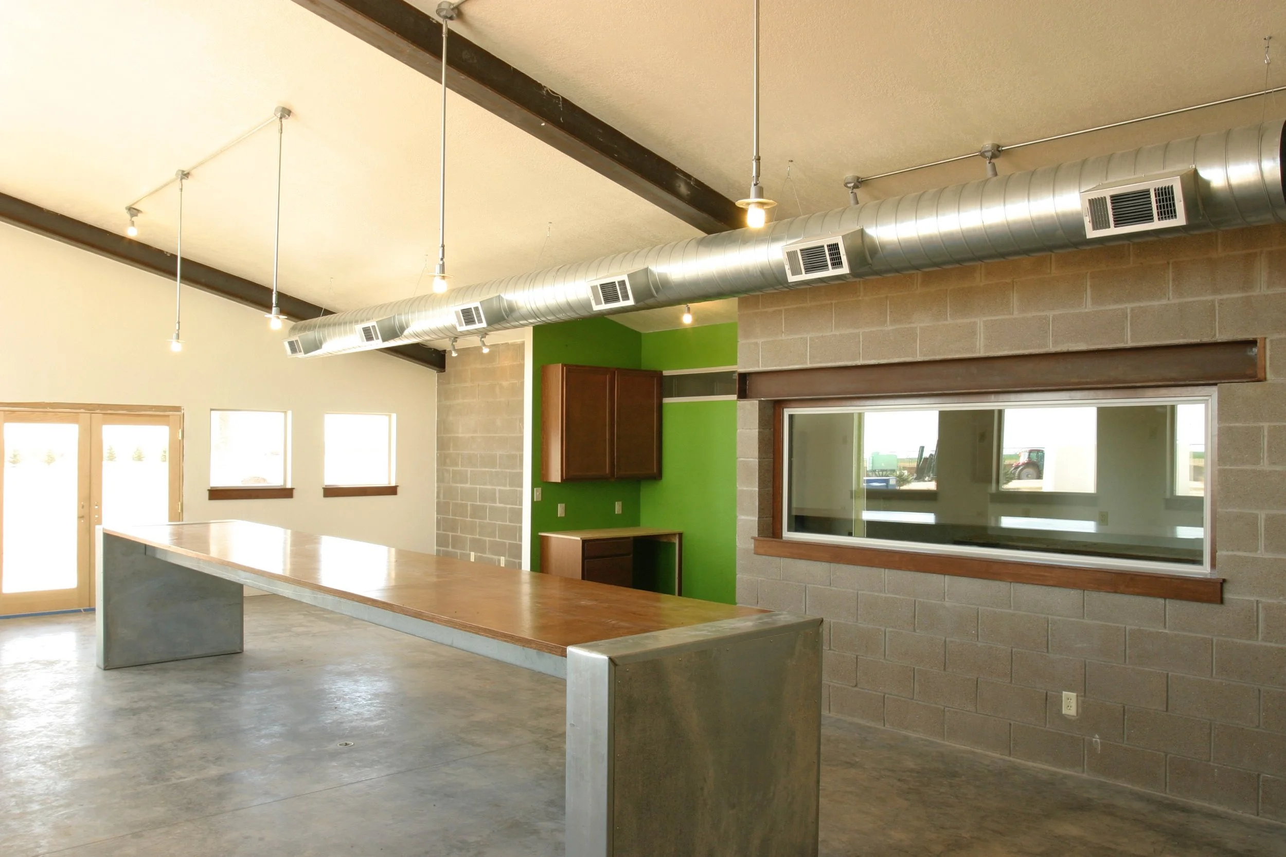 Interior of a modern commercial or industrial space with a long wooden table, exposed ductwork on the ceiling, green accent wall, and brick and concrete walls with windows and a door.