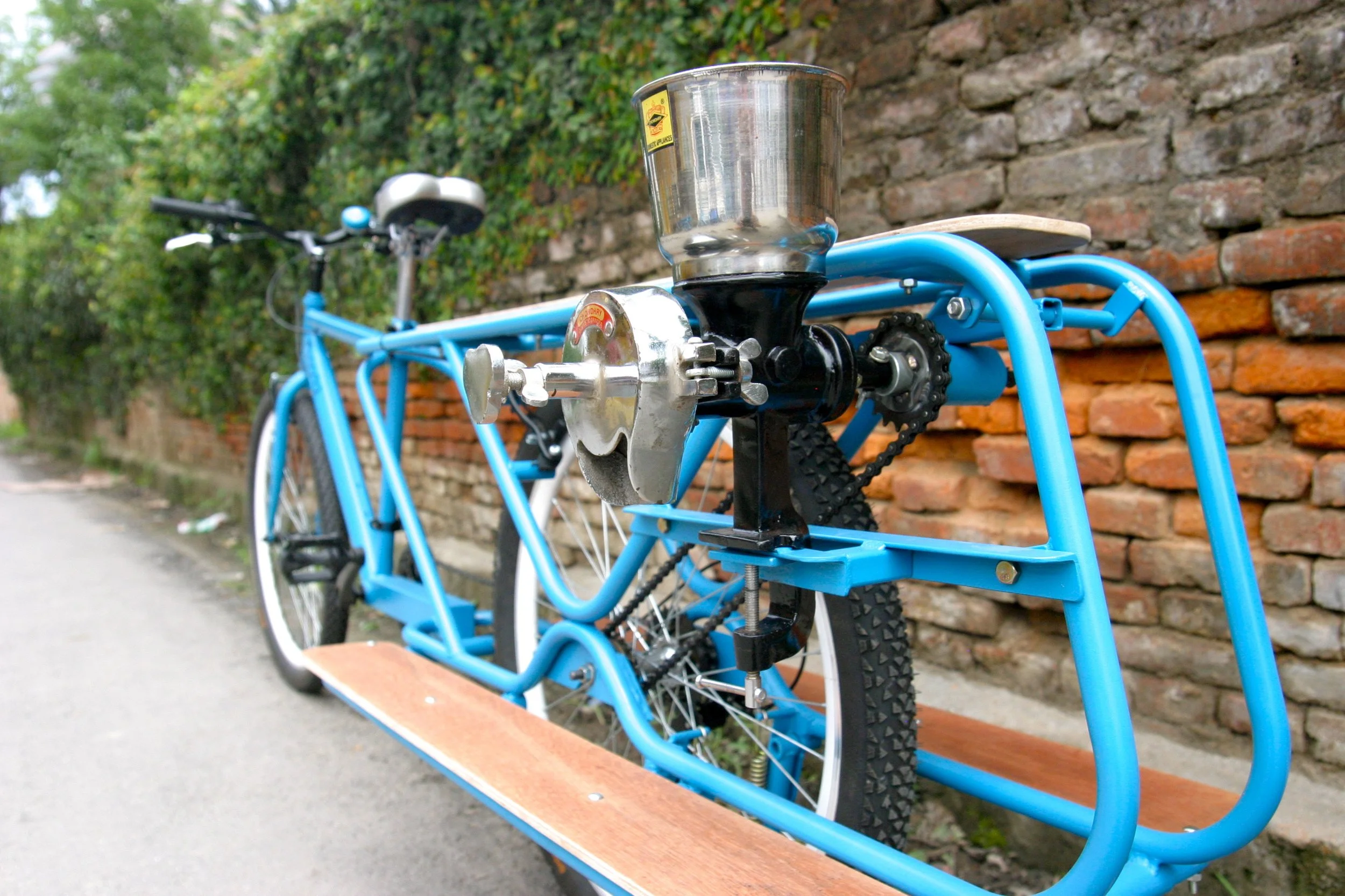 A blue bicycle converted into a stationary exercise bike with a grinding machine mounted on the frame, situated outdoors against a brick wall and greenery.