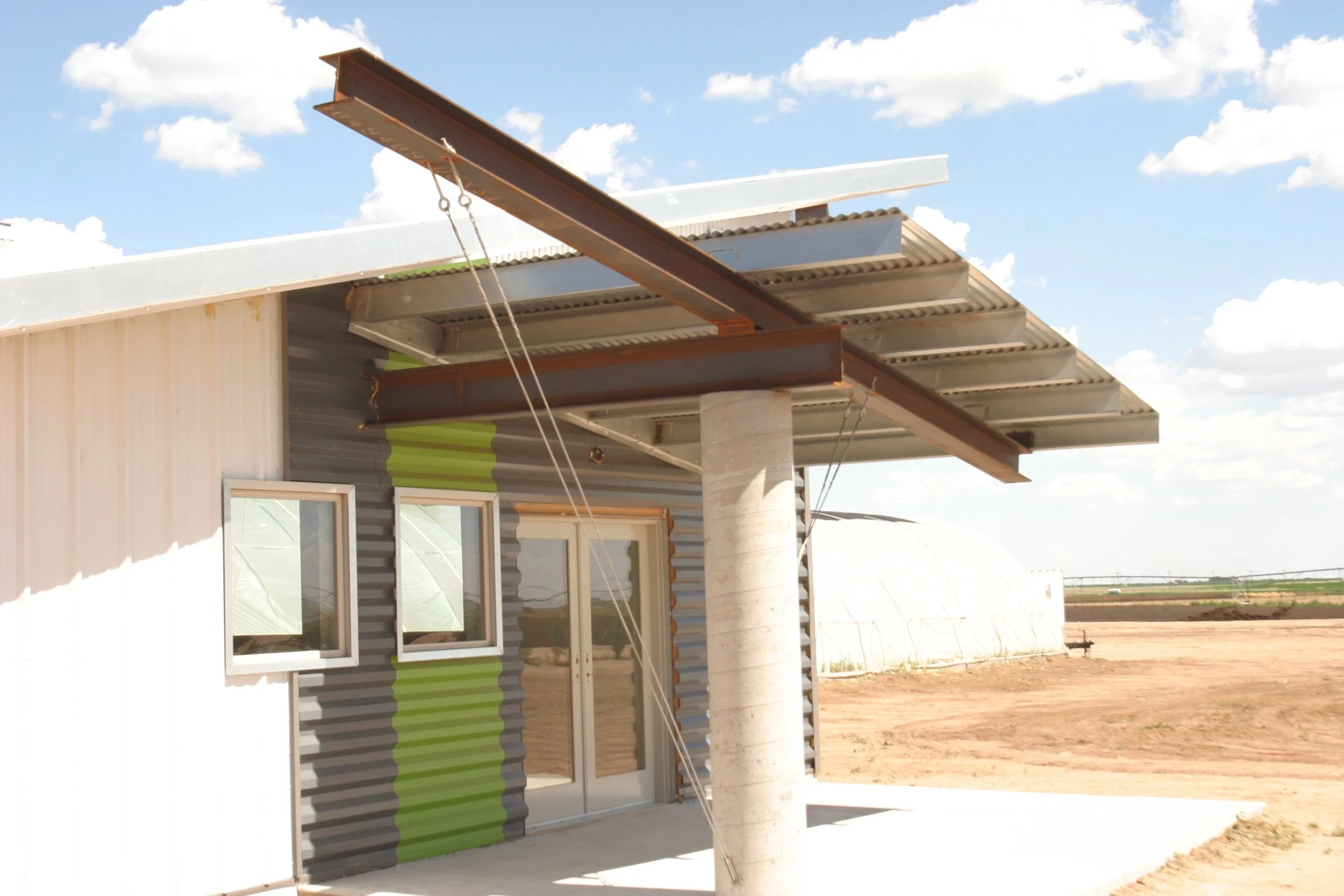 A building under construction with metal siding, two small windows, a glass door, and a cantilevered metal roof supported by a concrete column, set against a clear blue sky with scattered clouds.