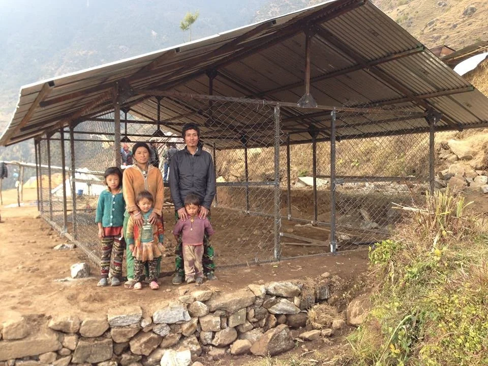 A family of five standing outside a chicken coop with a metal roof, surrounded by a chain-link fence, on a hillside in a rural area.
