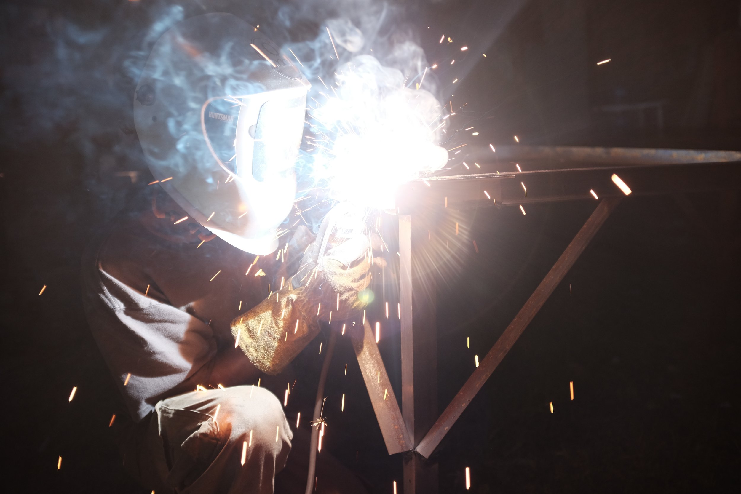 A person welding metal beams together outdoors at night, sparks flying from the welding process, wearing protective gear including a welding helmet and gloves.