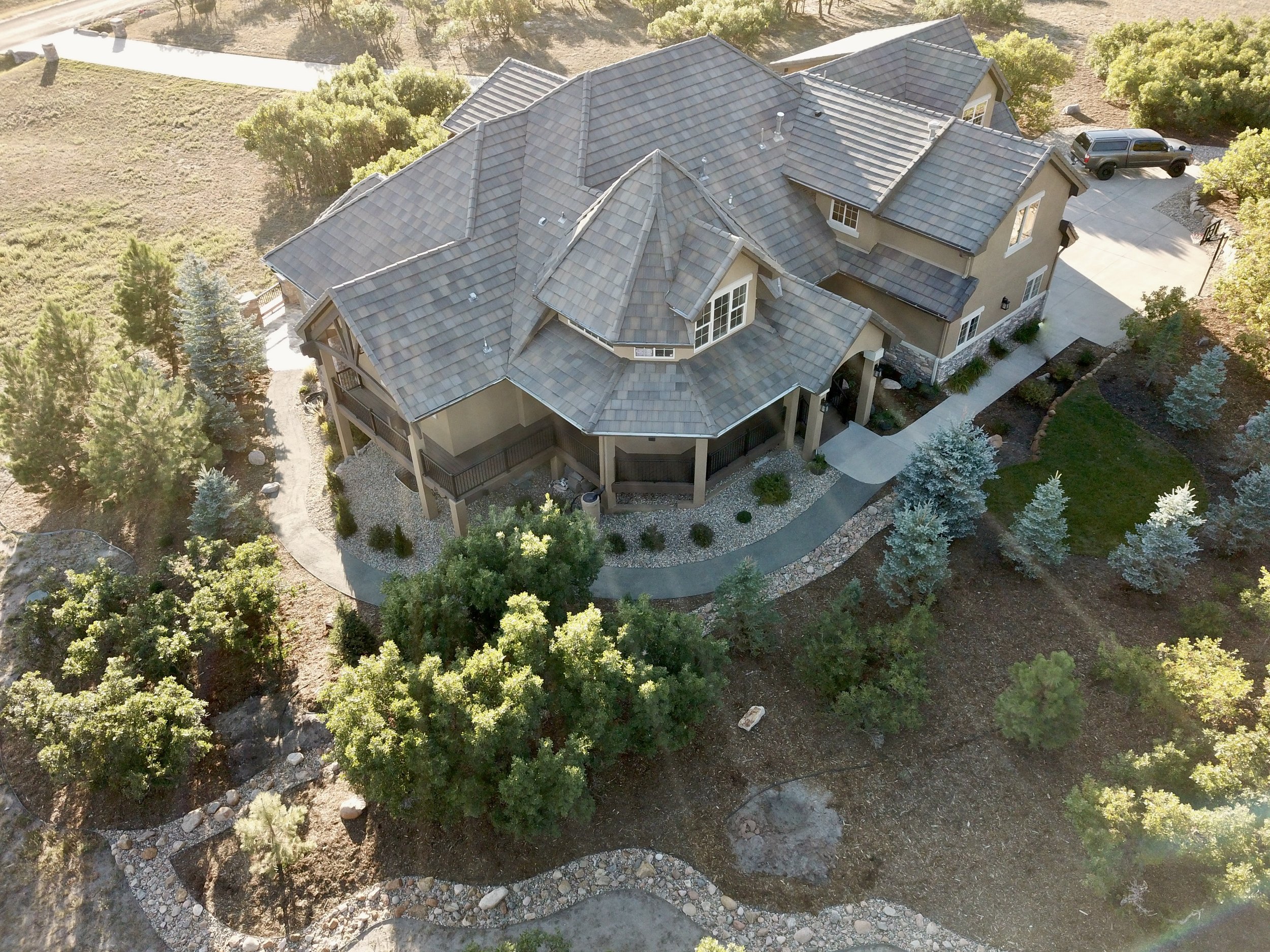 Aerial view of a large, multi-story house with a grey tiled roof, surrounded by landscaping, trees, bushes, and a driveway with parked vehicle.