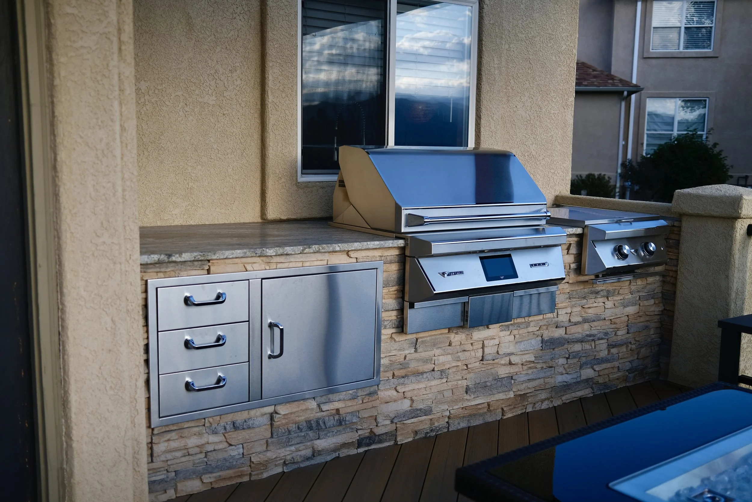 Outdoor kitchen with stainless steel grill, side burners, and storage cabinets mounted on a stone wall.