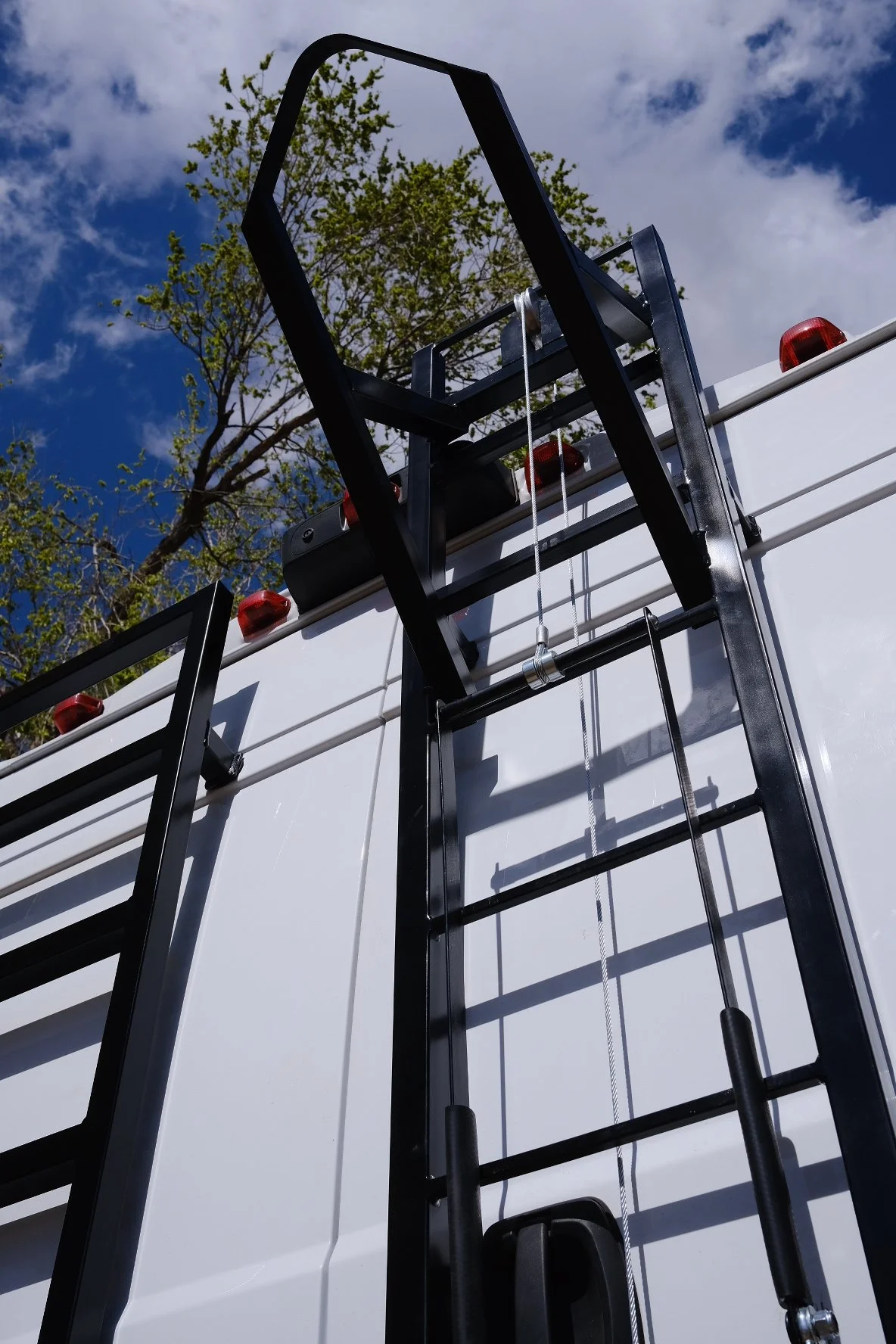 The image shows a close-up of a fire truck's ladder and aerial platform against a background of a partly cloudy sky and green trees.