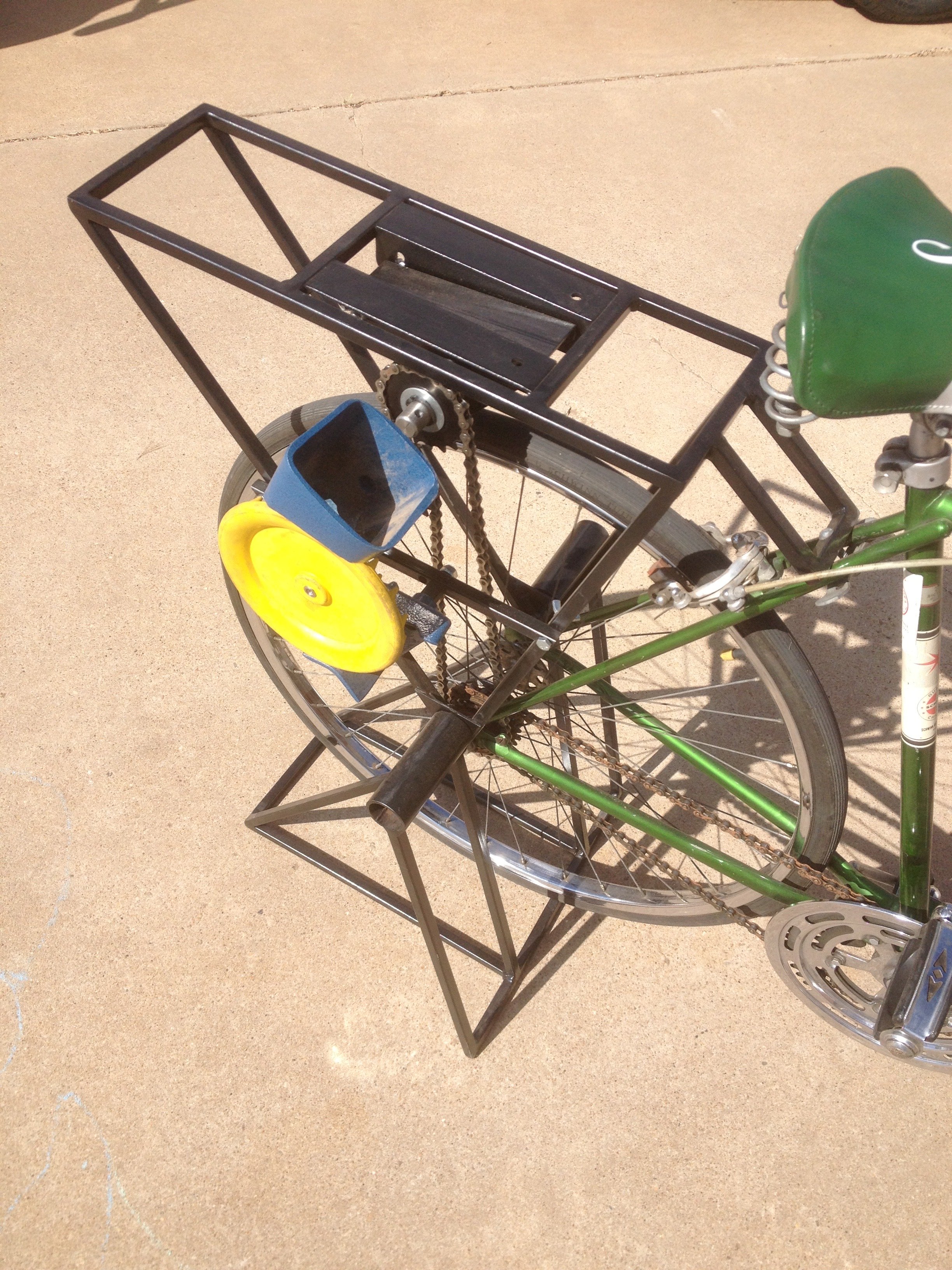A bicycle with a black metal frame carrier on the back, a green seat, and blue and yellow front wheels, parked on a concrete surface.