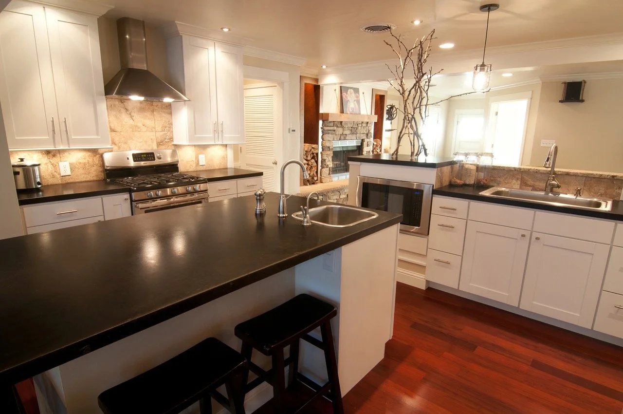 Modern kitchen with white cabinets, black countertops, stainless steel appliances, and hardwood floors. There is a kitchen island with a sink and seating, a stone fireplace in the background, and a decorative tree branch centerpiece.