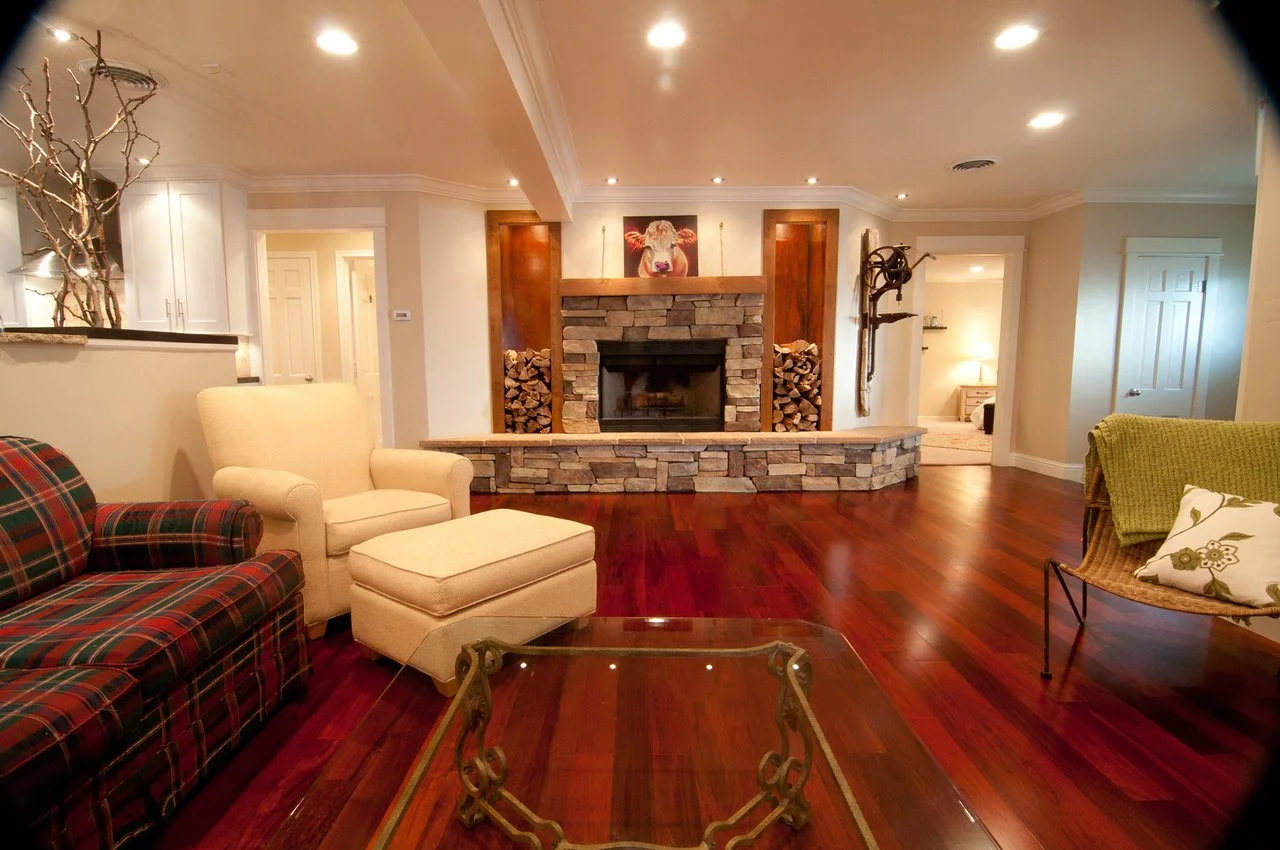 Living room with wood floor, fireplace with stone surround, and furniture including a plaid sofa, cream armchair with ottoman, and wicker chair with a green blanket. There is a glass coffee table in the center, and a painting of a cow above the firep
