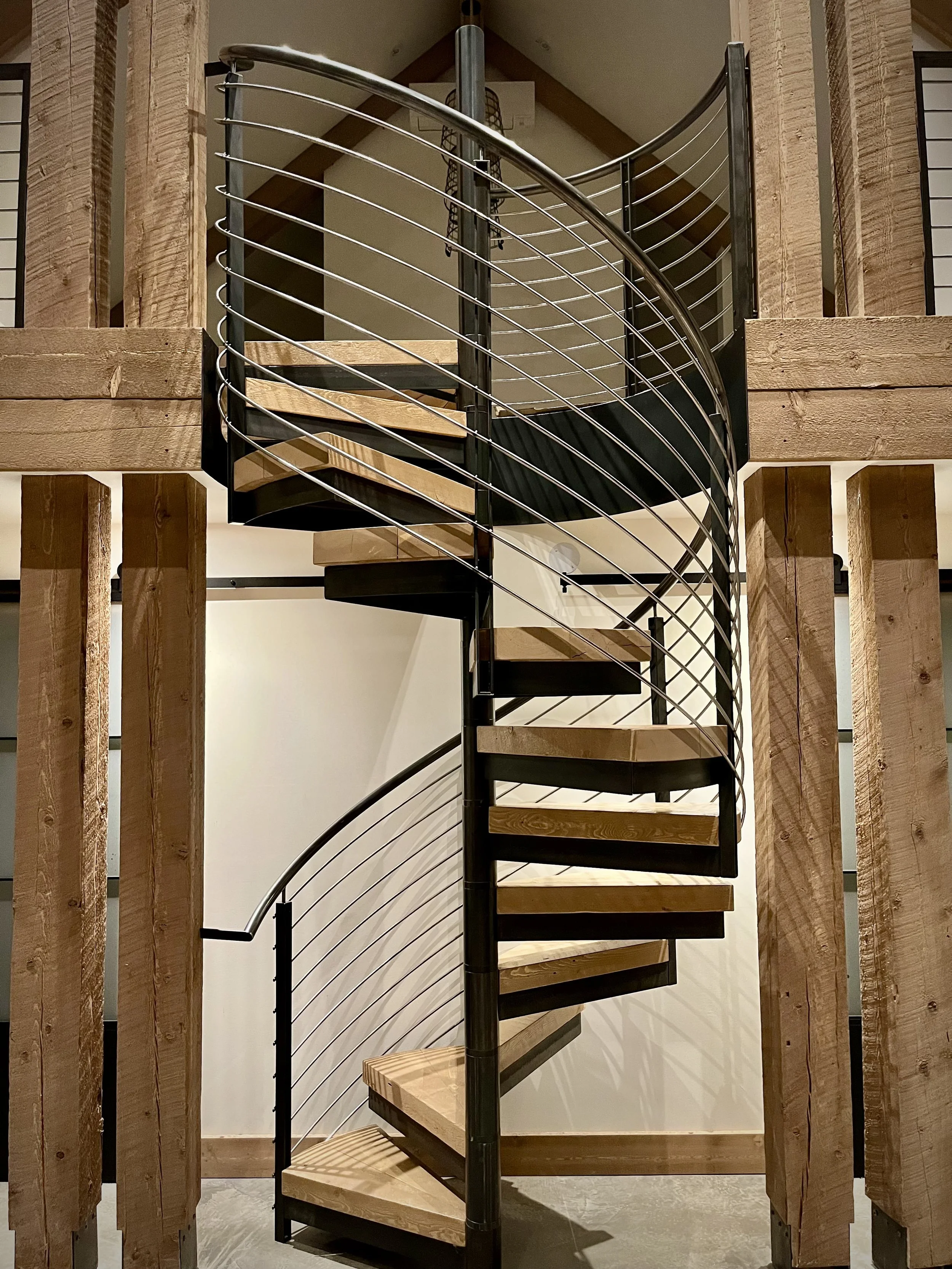 A spiral staircase with wooden steps, black metal supports, and horizontal metal railings, situated between wooden beams in an interior space.
