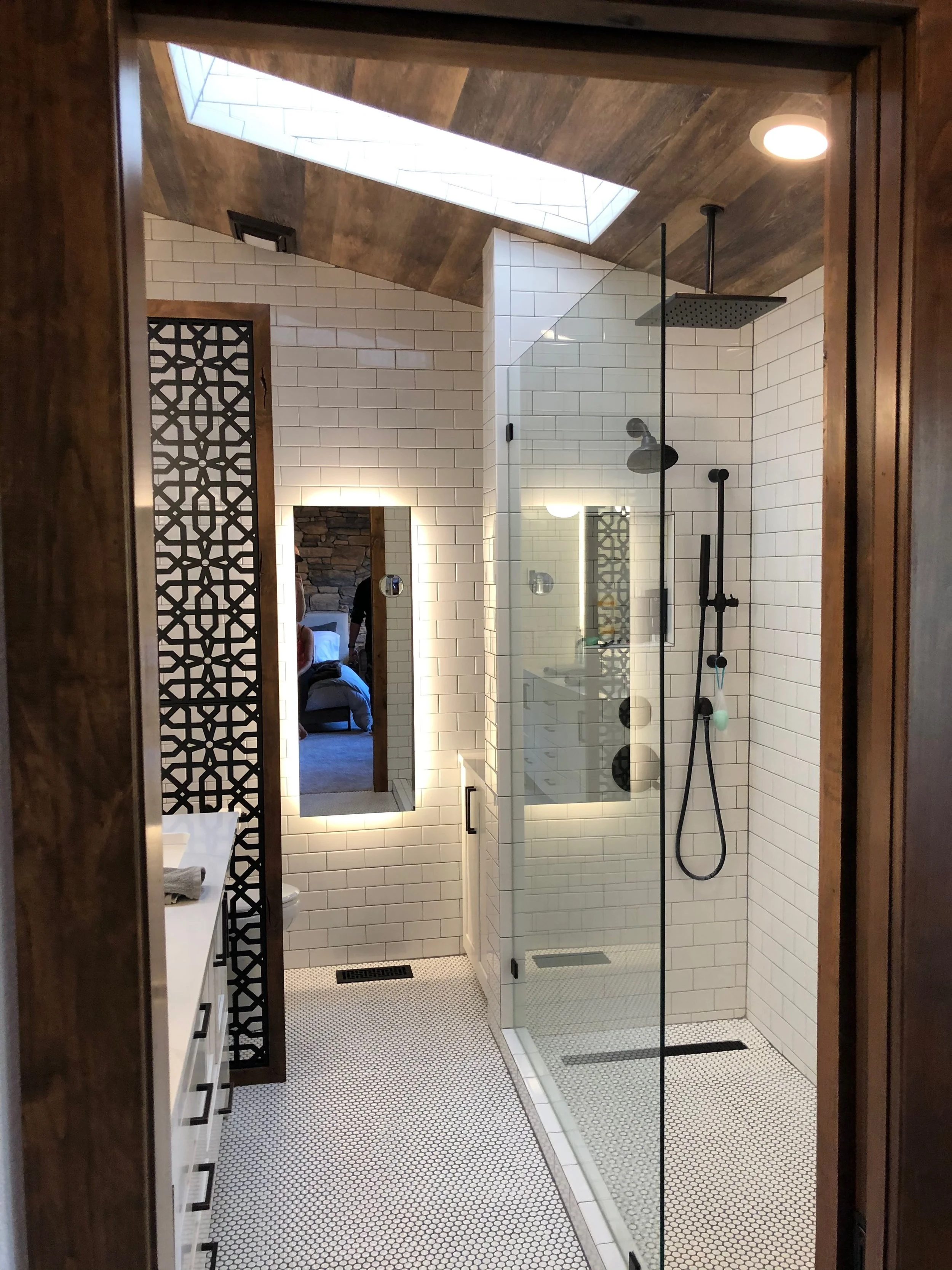 A modern bathroom with a glass-enclosed shower, white subway tile walls, a skylight, a mirror with backlighting, and a patterned black decorative room divider.