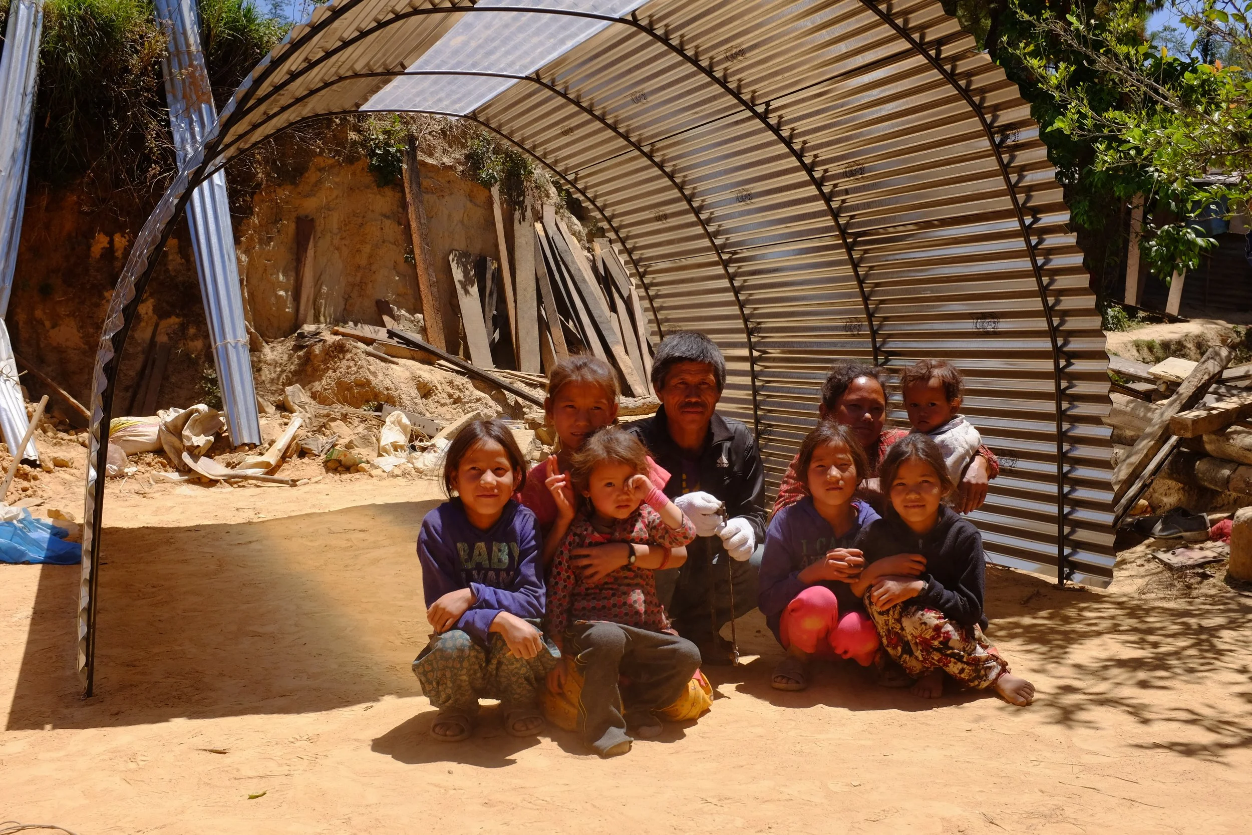 Group of children and a man posing inside a metal greenhouse structure on dirt ground, with construction debris and rocky terrain behind them.