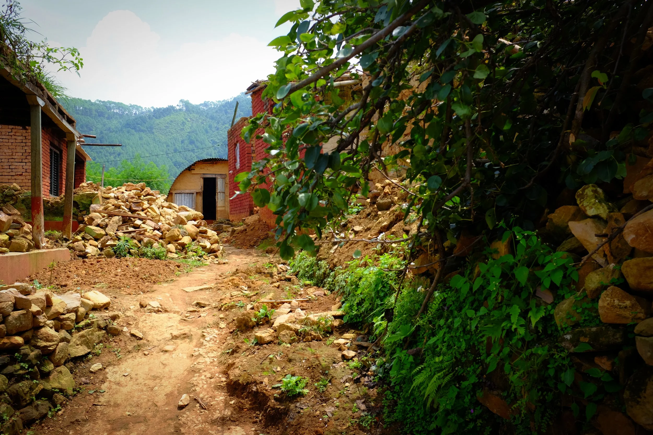 A dirt path in a rural village with stone walls, brick houses, and greenery, with mountains in the background.