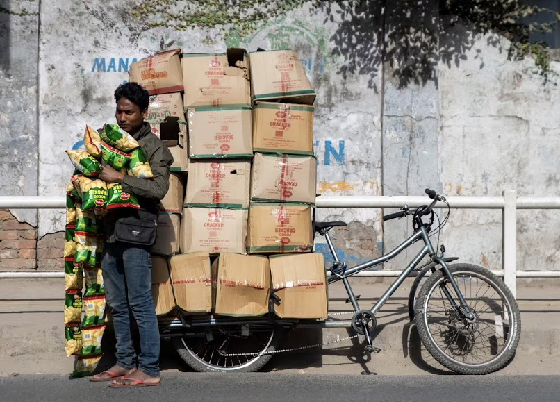A man standing on the street near a bicycle loaded with cardboard boxes and snack bags.