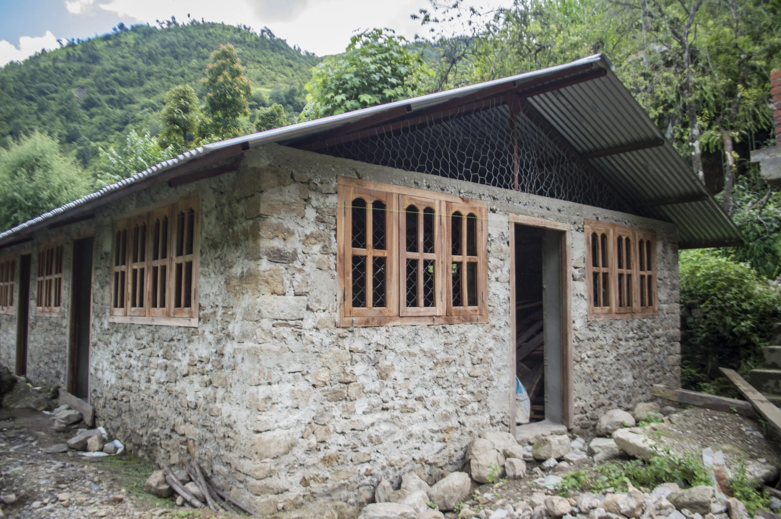 A stone house with wooden window frames and a metal roof, situated in a green, hilly landscape.