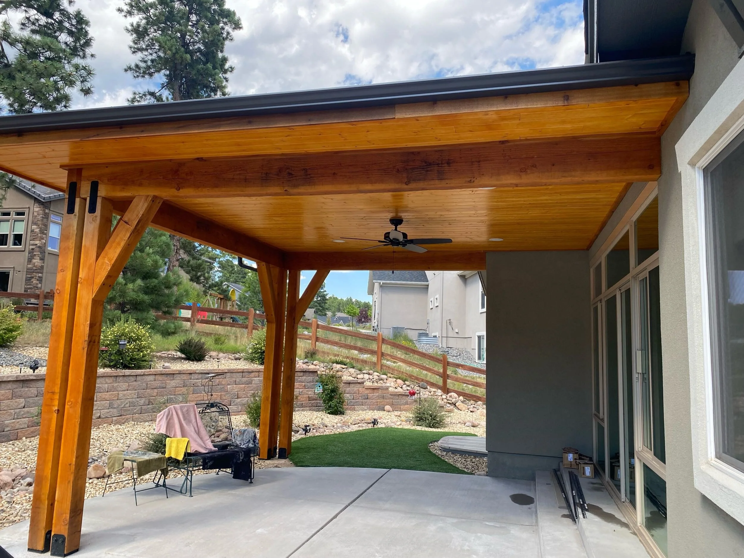 Back porch with a wooden ceiling, a ceiling fan, and a concrete floor, with some outdoor furniture, boxes, and tools, overlooking a landscaped yard with trees and a wooden fence.