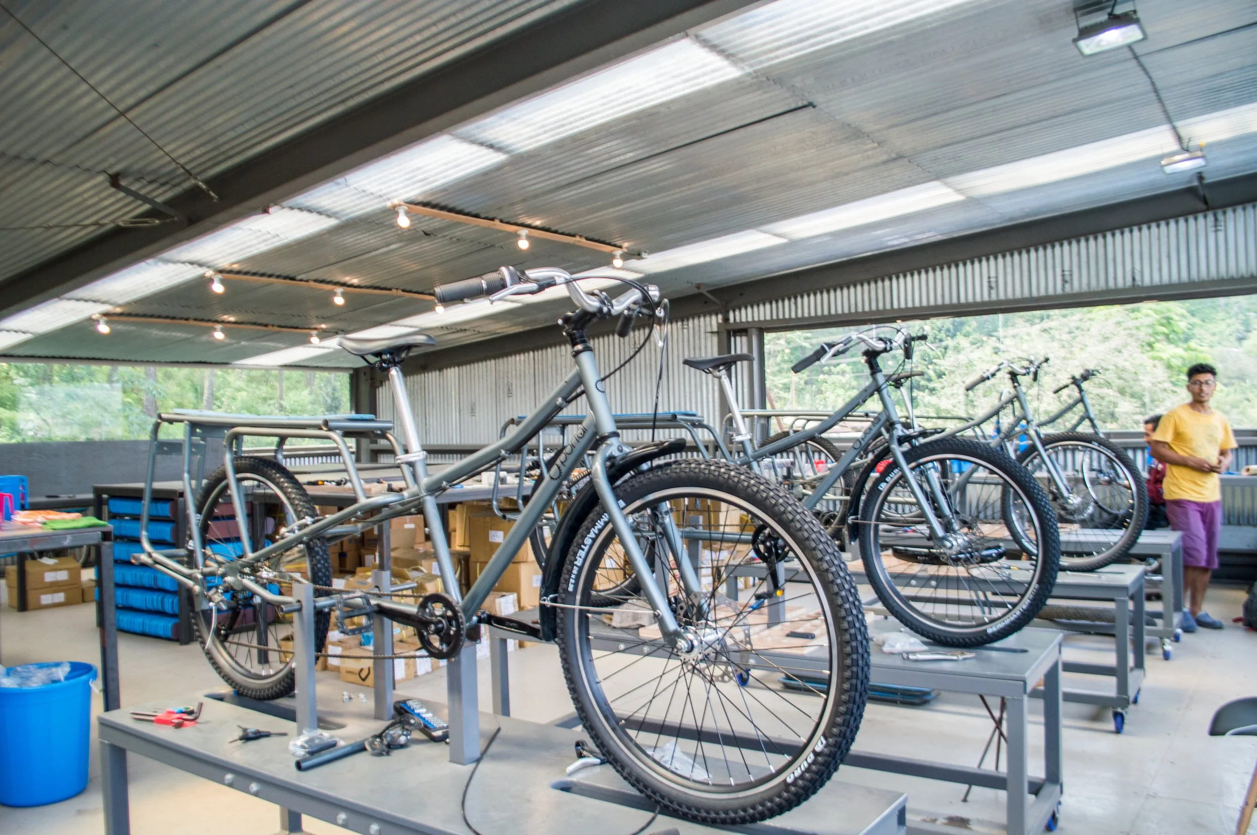 Indoor workshop with several dark gray bicycles on workbenches, with tools and boxes around, and people in the background.