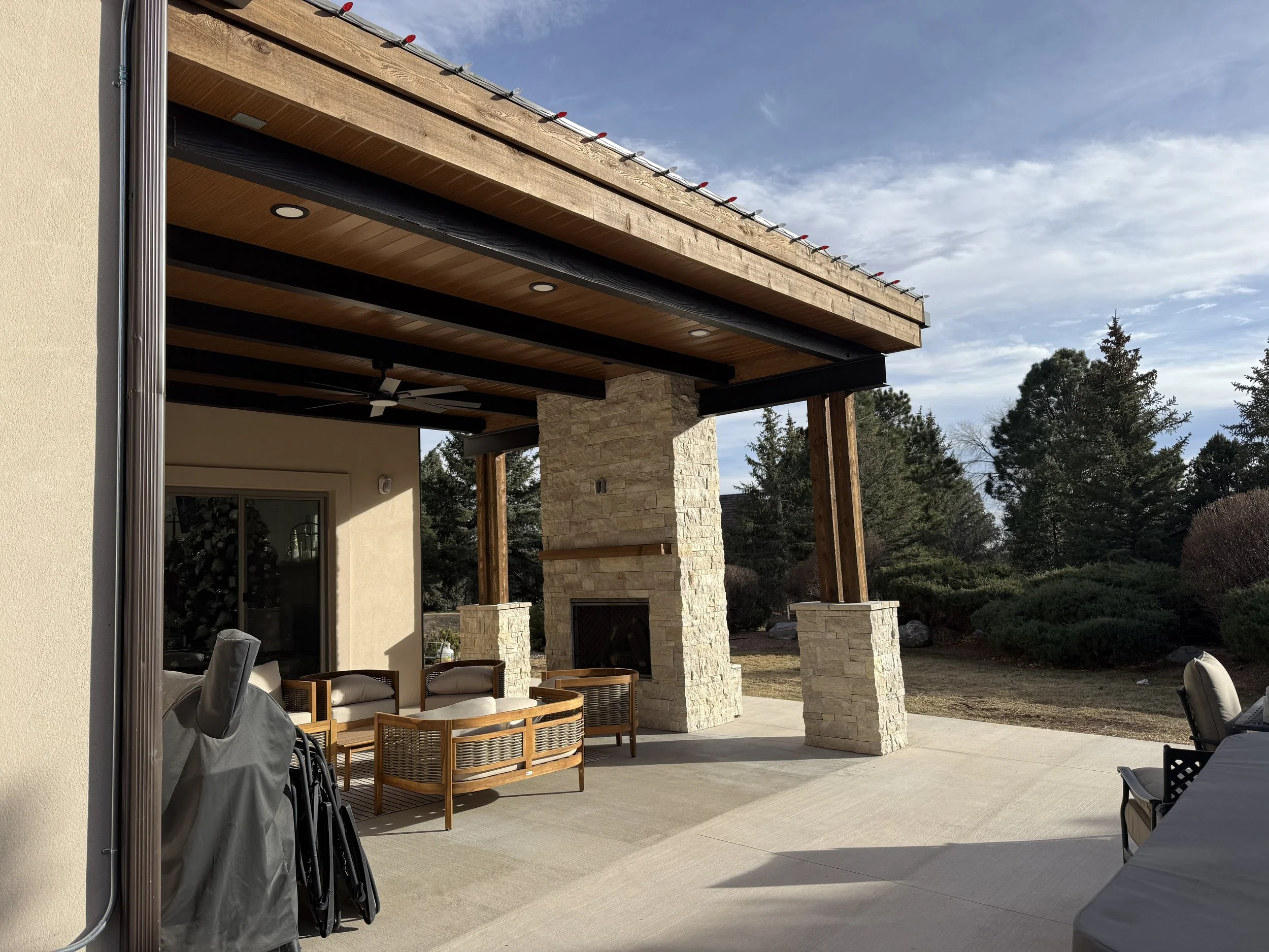 Outdoor patio with seating area, stone fireplace, and covered roof, with trees and sky in the background.